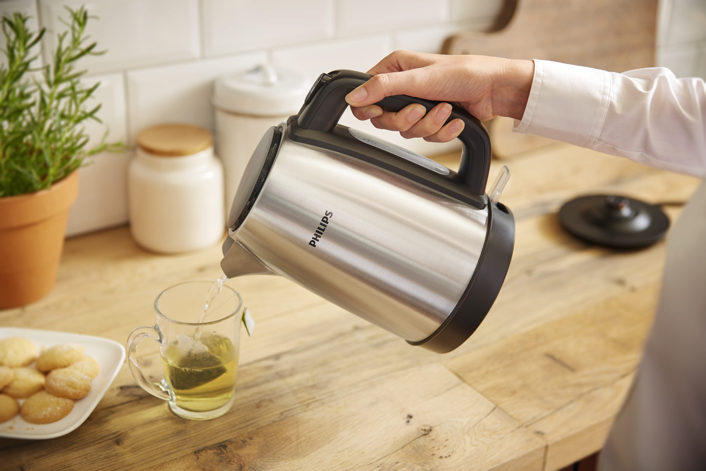 Person gießt Wasser aus einem silbernen Philips-Wasserkocher in eine Teetasse. Kekse auf Holztisch.