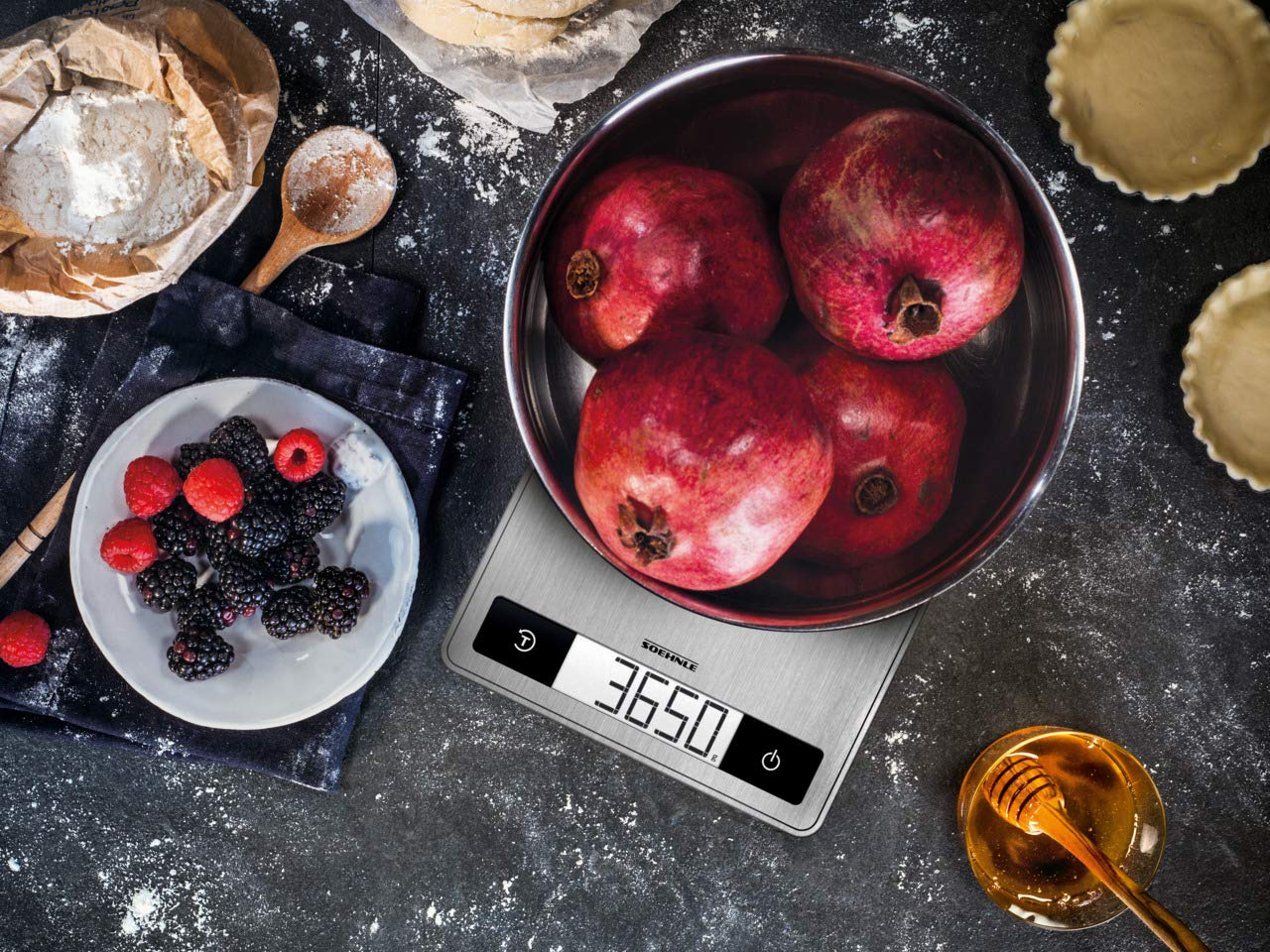 Eine Waage mit Granatäpfeln, Beeren und Honig. Anzeige zeigt 365,0. Dunkler Hintergrund.