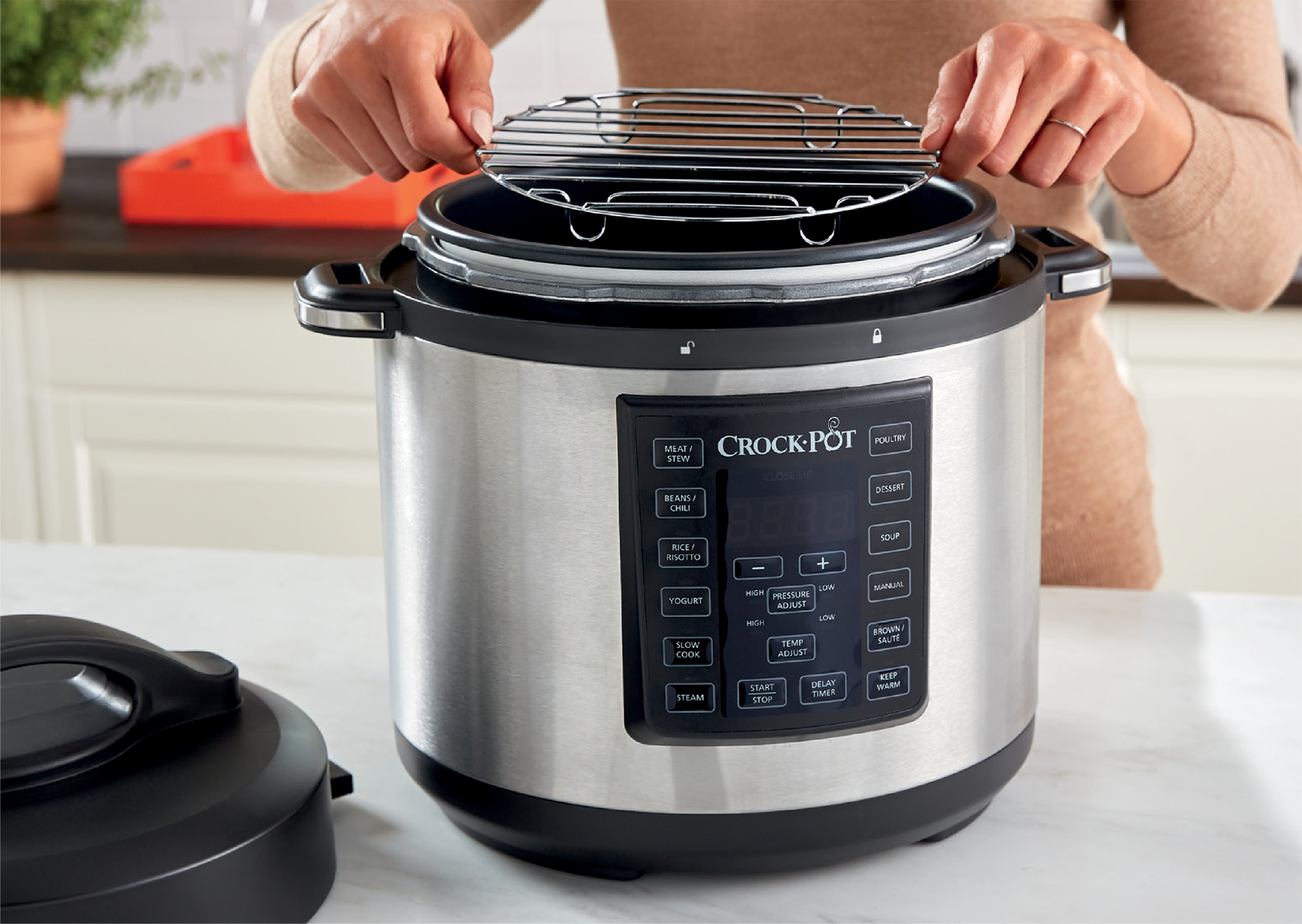 Handen die een metalen rek boven een zilveren Crock-Pot slowcooker houden. Bedieningspaneel zichtbaar.