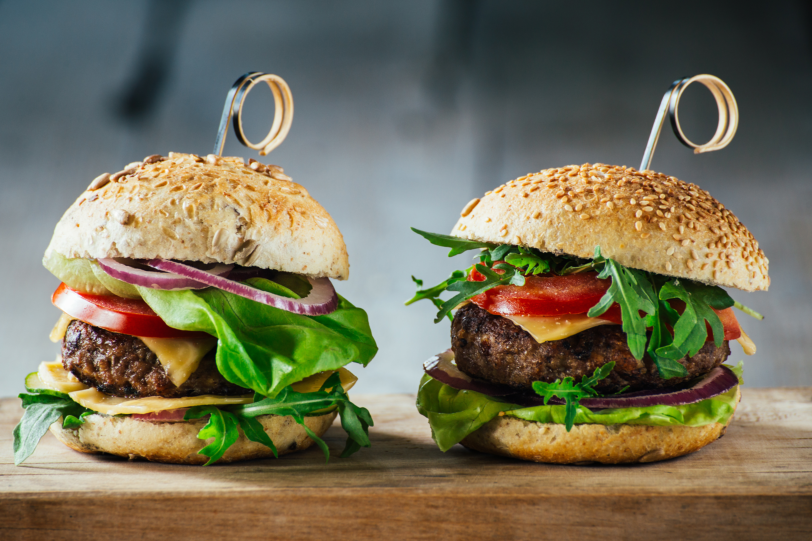 Zwei Burger auf einem Holzbrett. Sie haben Sesambrötchen und sind mit Belägen gefüllt.