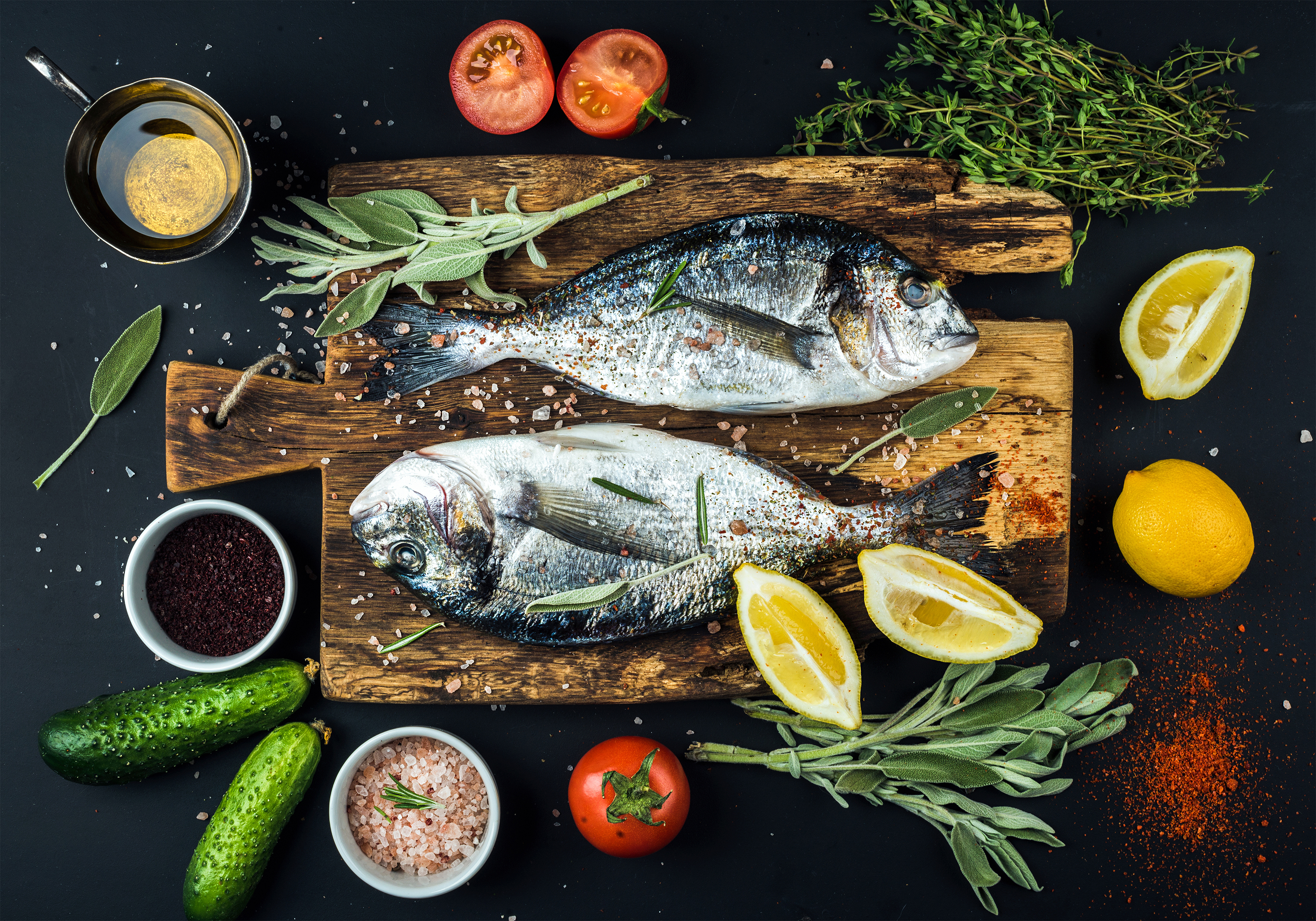 Zwei Fische auf einem Holzbrett mit Zitronen, Gurken und Gewürzen auf schwarzem Grund.