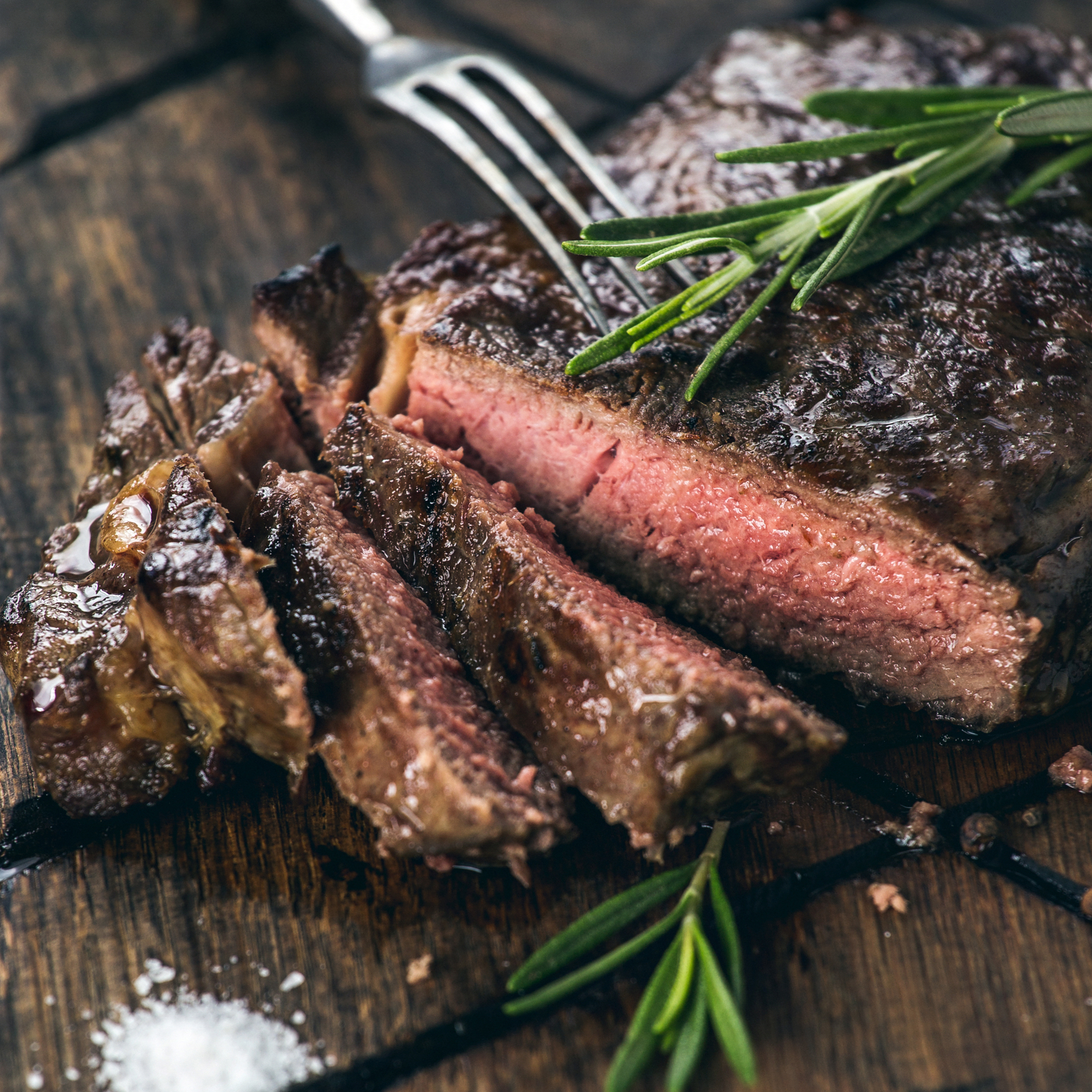 Geschnittenes Steak auf einem Holzbrett mit Rosmarin und Salz, eine Gabel steckt im Steak.