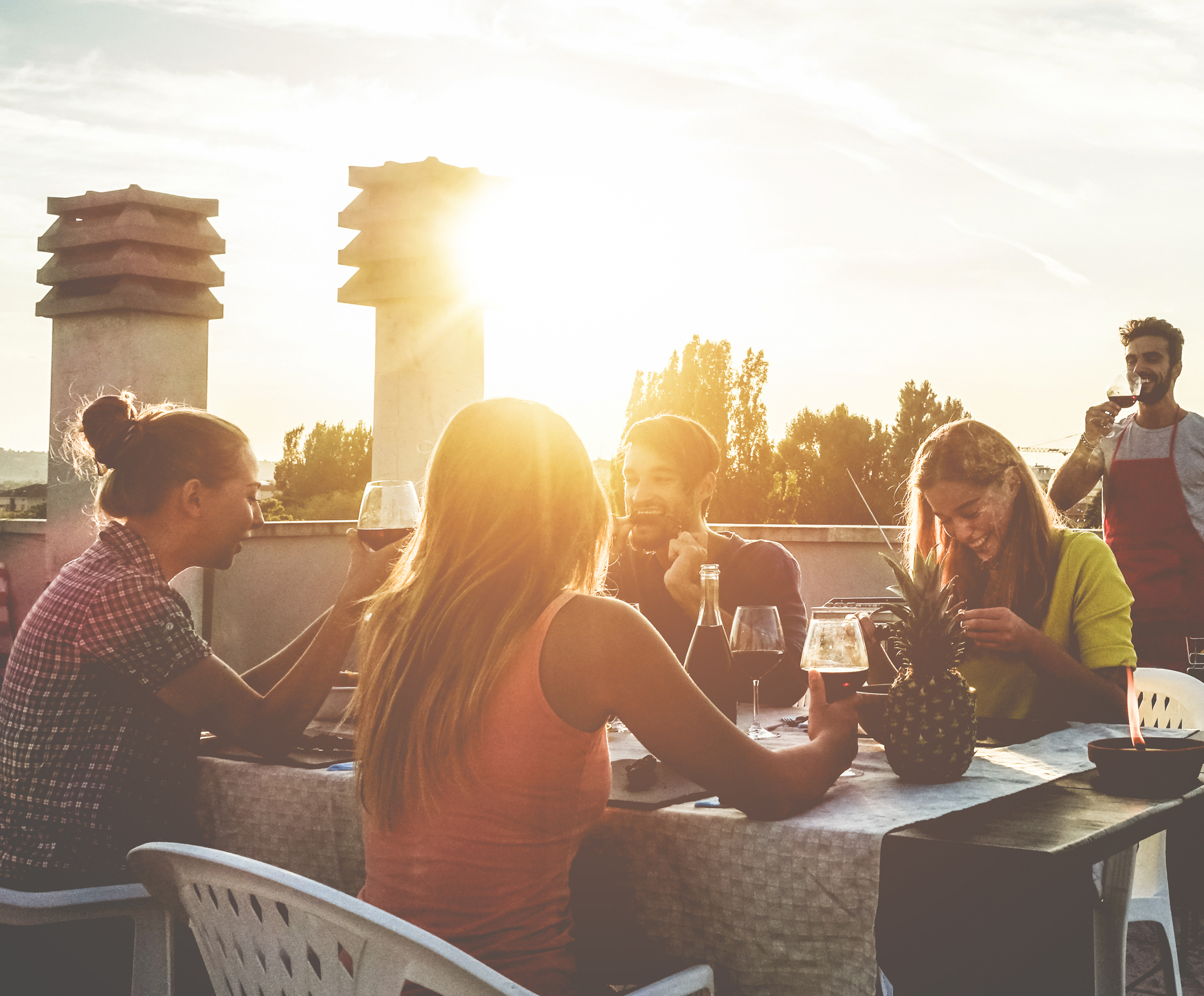 Eine Gruppe von Leuten genießt Getränke und eine Mahlzeit auf einer Dachterrasse bei Sonnenuntergang. Eine Ananas steht auf dem Tisch.