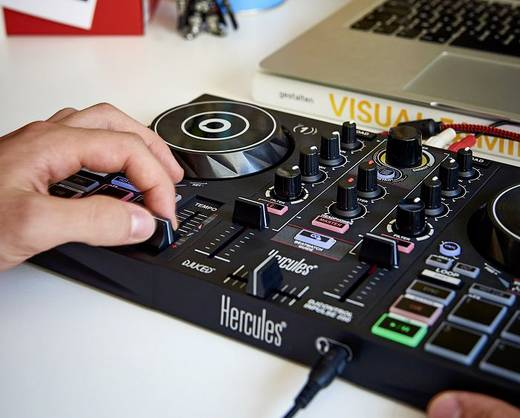 La mano de una persona ajustando una mesa de mezclas de DJ, con un portátil al fondo.