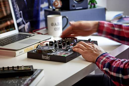 Manos de una persona sobre un controlador de DJ. Una computadora portátil está abierta en el fondo. Taza blanca: "Game On".