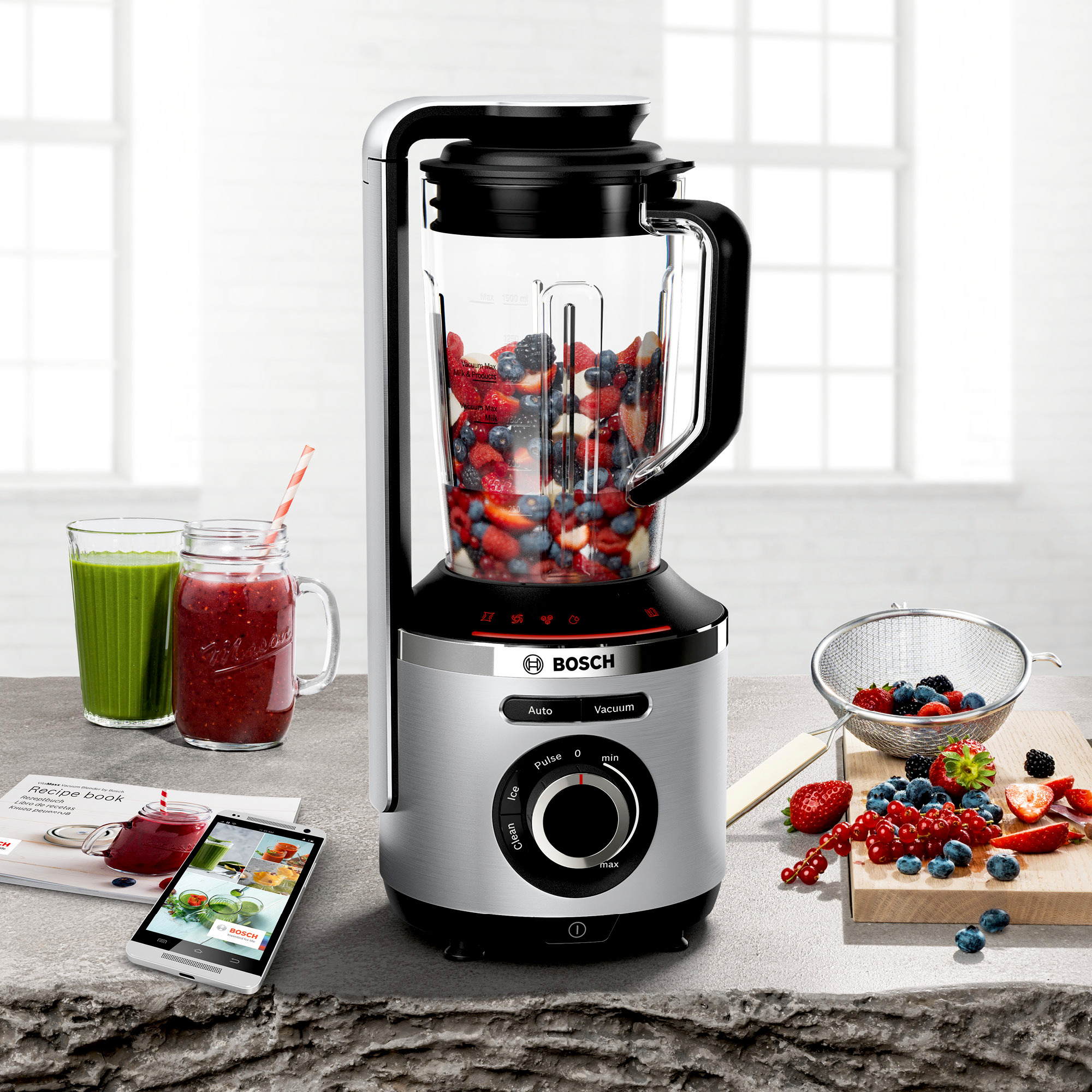 Ein silberner Mixer mit einem Glaskrug, gefüllt mit gemischten Früchten, neben Obst und einem Rezeptbuch.