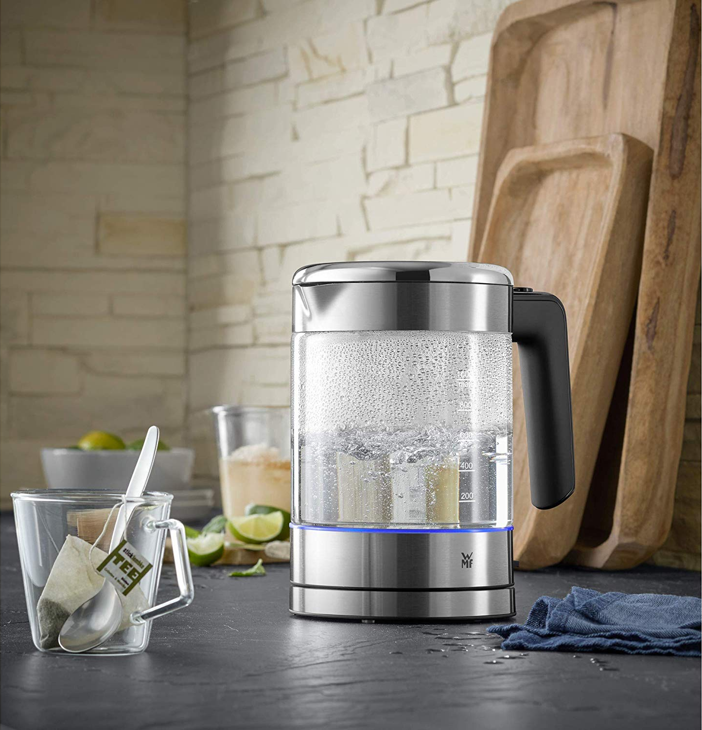 Bollitore elettrico in vetro con acqua bollente, una tazza con bustina di tè e vassoi di legno sullo sfondo.