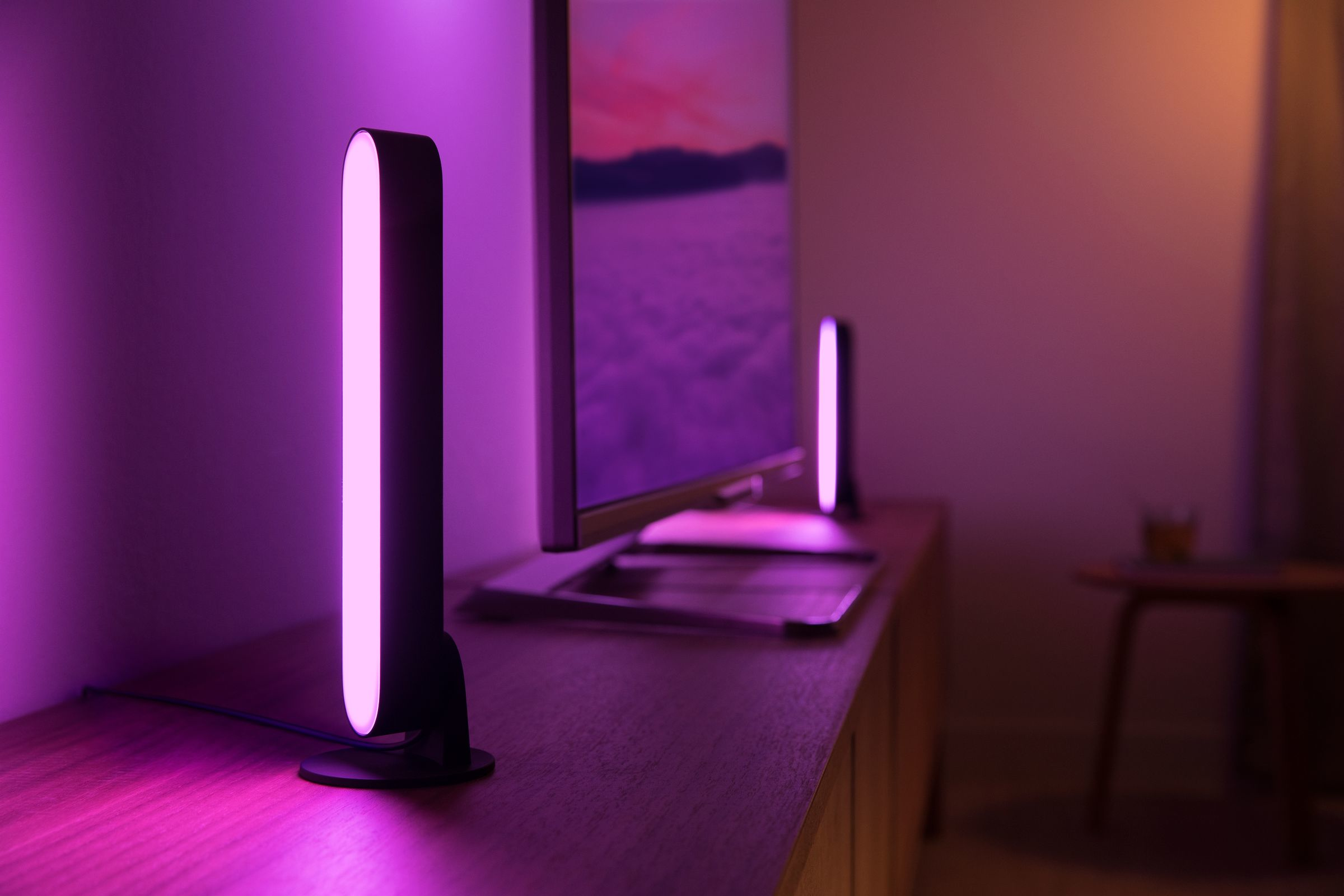 Due luci viola verticali su un tavolo di legno, con una TV sullo sfondo. Illuminazione viola.
