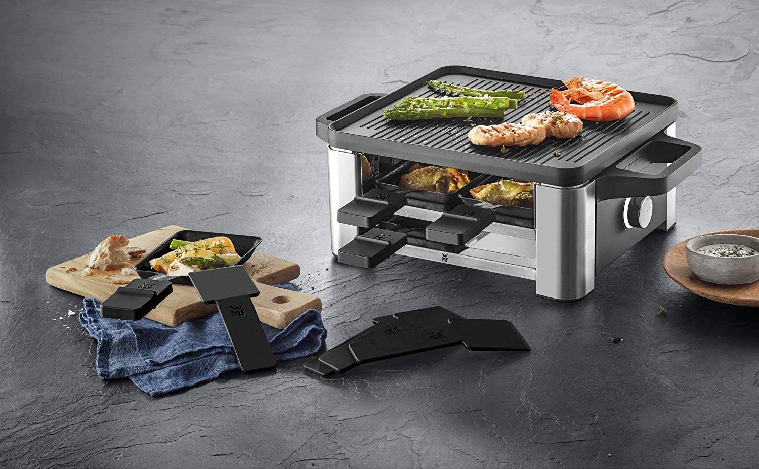 Raclette-Grill mit Essen oben und im Grill unten, mit Servierzubehör.