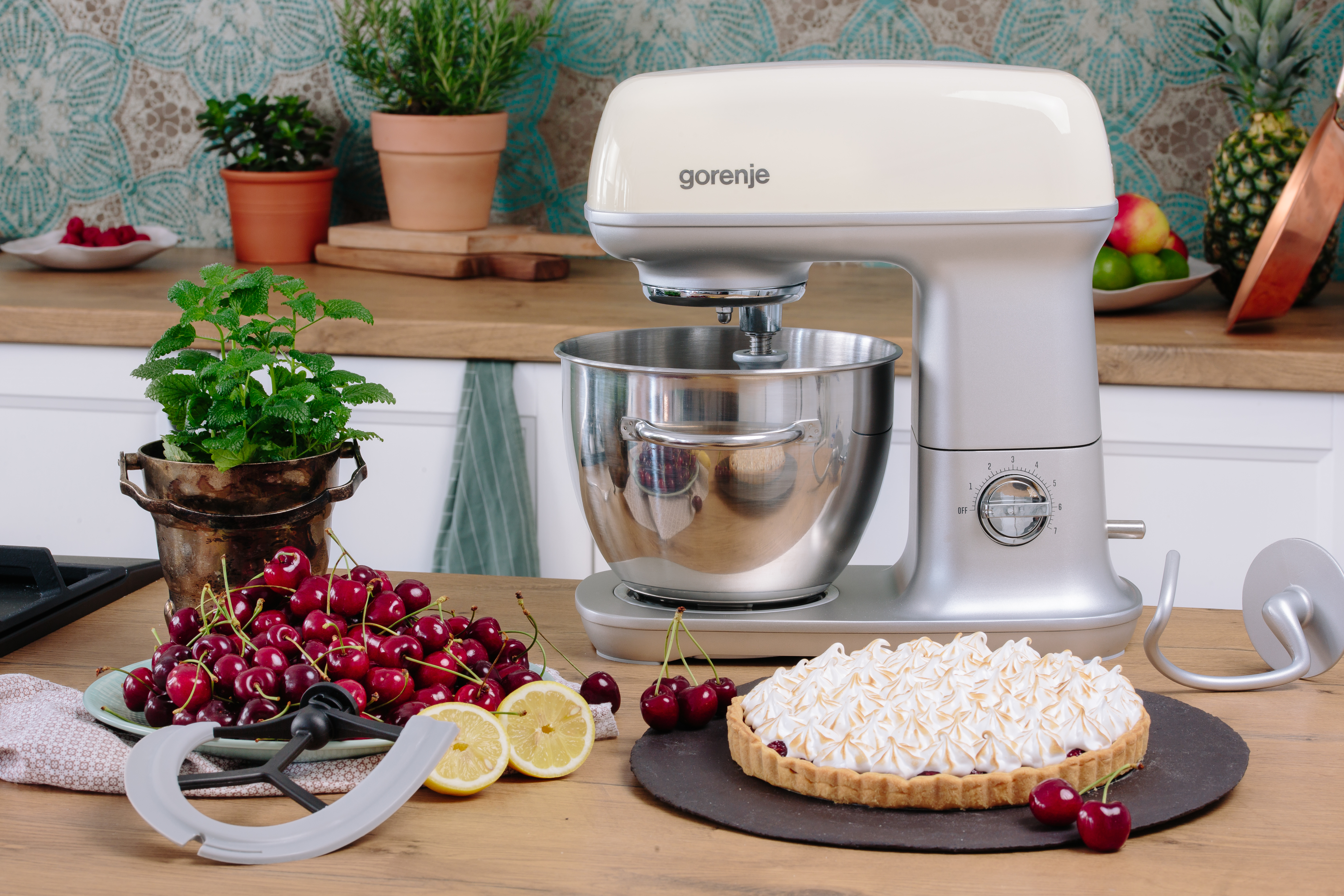 Egy konyhai mixer egy asztalon áll, cseresznyés pitével és cseresznyével. Egy vágódeszka és citromok vannak az asztalon.