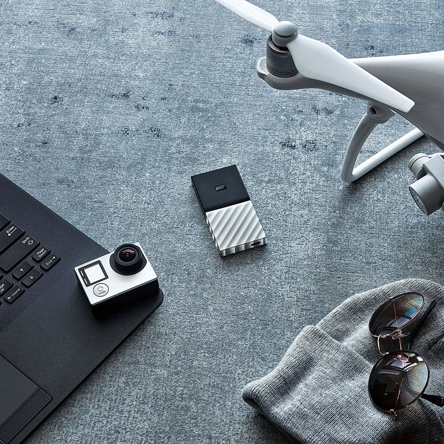 Laptop, camera, external hard drive, drone, beanie, and sunglasses on a grey surface.