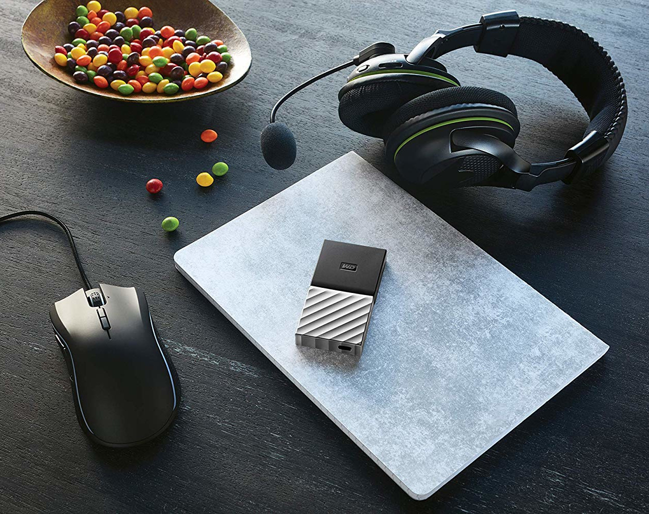 A laptop, headphones, mouse, external hard drive, and a bowl of colorful candies on a black surface.