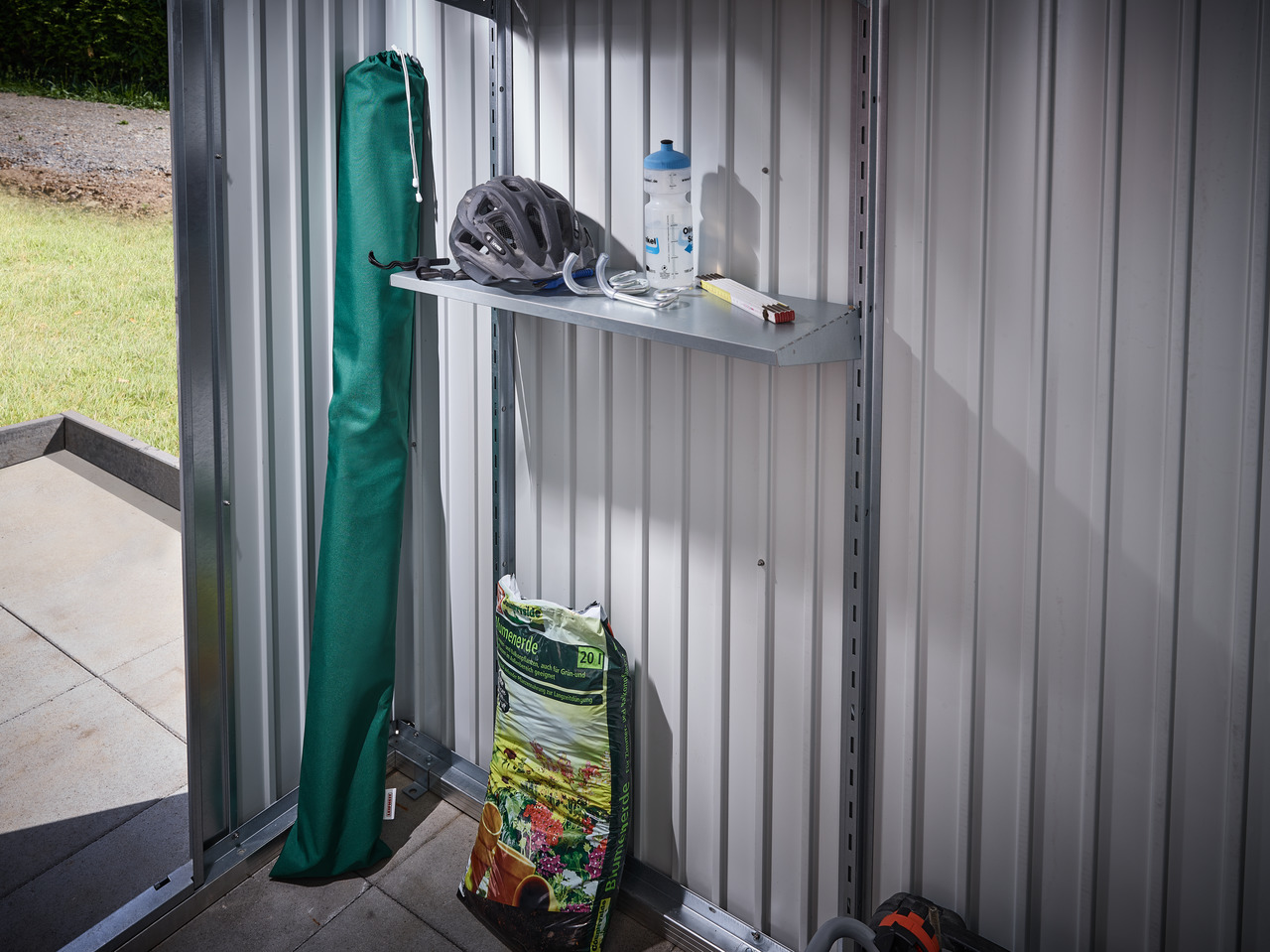 Ein offener Metallschuppen mit einem Regal. Grüne Tasche, Fahrradhelm, Wasserflasche und andere Gegenstände sind vorhanden.