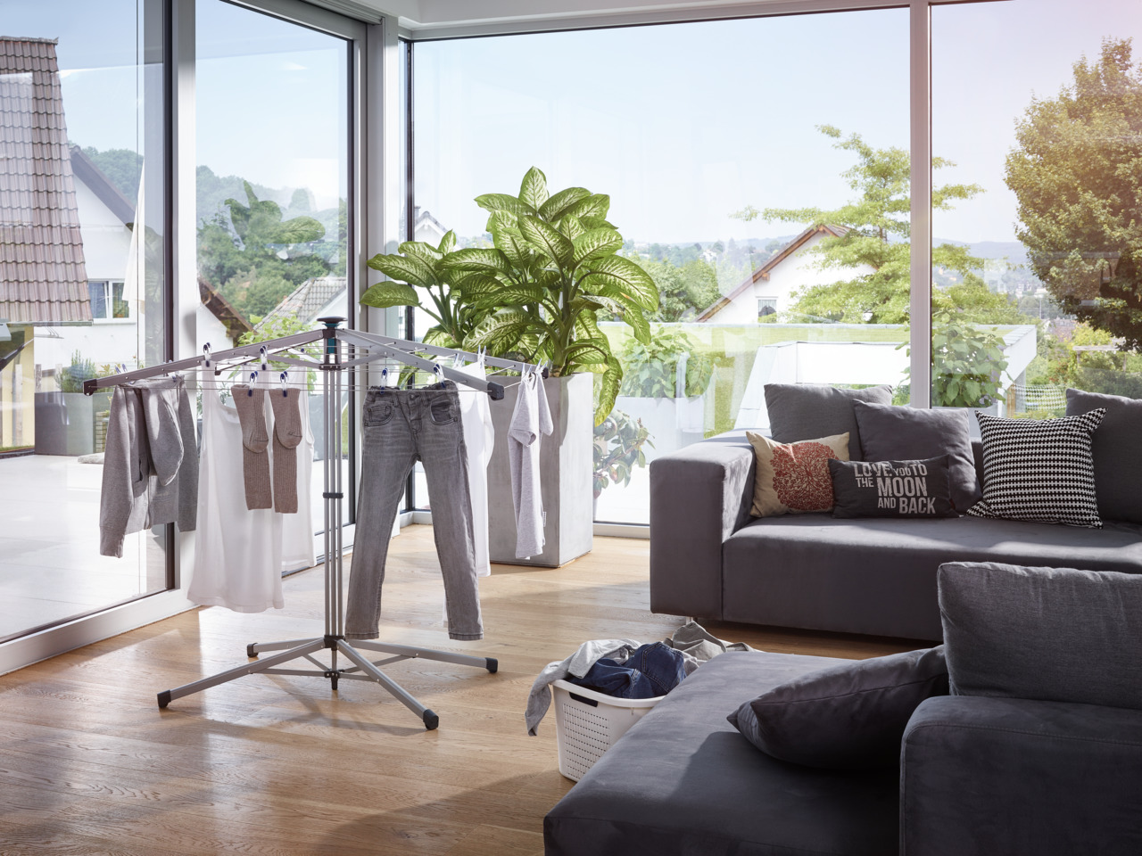 Wäscheständer mit Kleidung, in einem Raum. Sofa, Pflanzen und Fenster im Hintergrund.