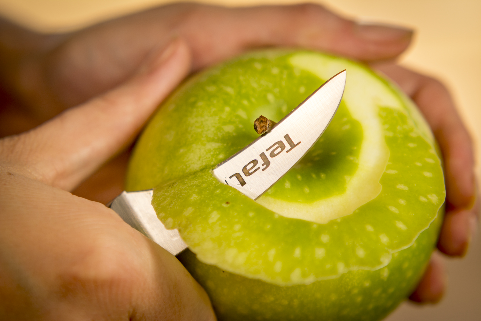 Kéz zöld almát hámoz késsel, a késsel a Tefal szó van nyomtatva.