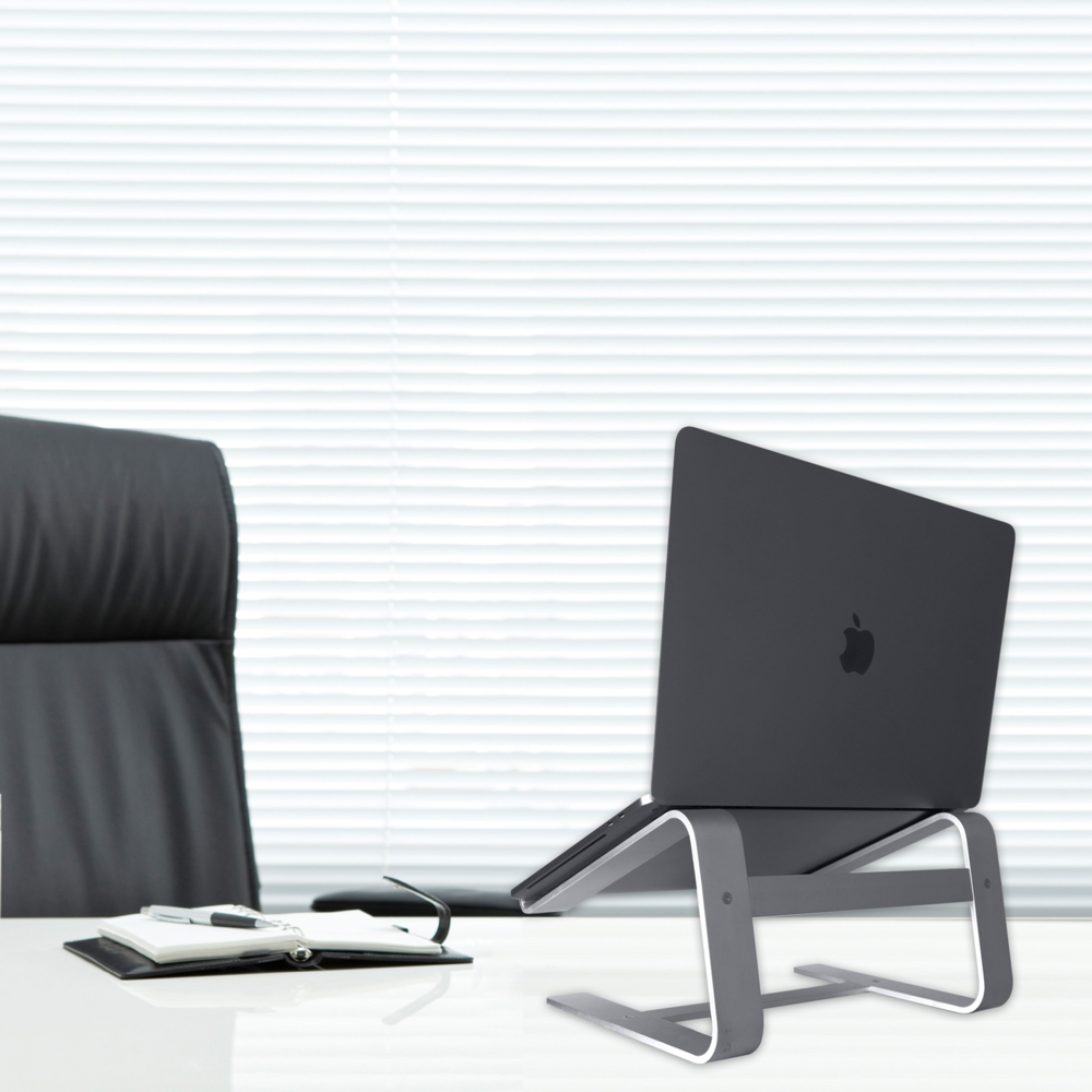 Ordinateur portable sur support argenté, sur un bureau blanc, avec une chaise noire et des stores derrière.