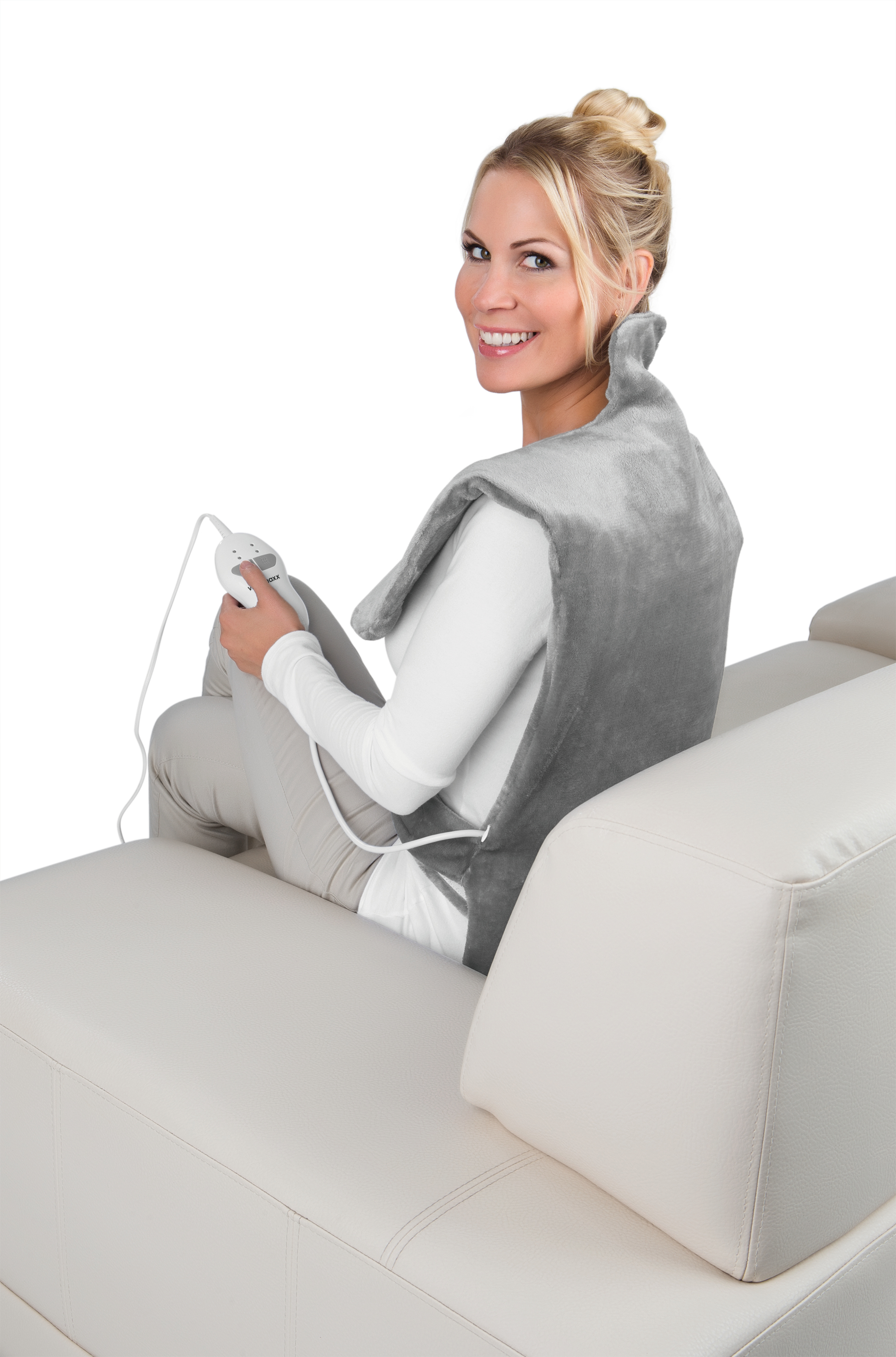 Frau sitzt auf einem weißen Sofa mit einem grauen Heizkissen auf dem Rücken und lächelt in die Kamera.