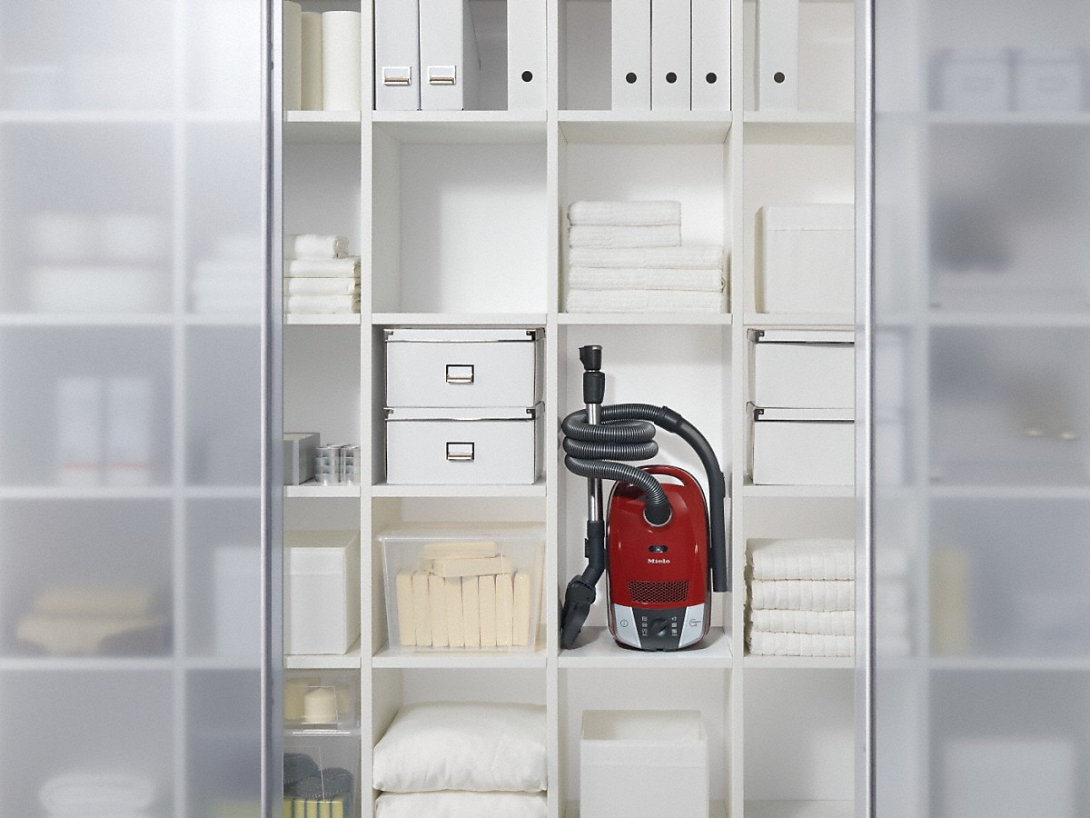 Ein weißer Schrank mit Regalen. Ein roter Staubsauger steht in der Mitte.