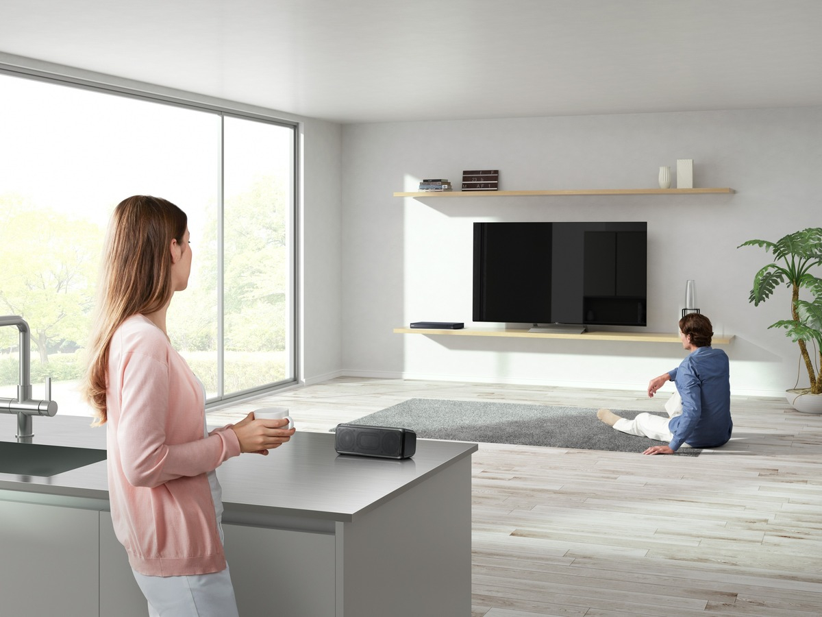 Frau mit Tasse blickt auf einen Mann, der in einem modernen Wohnzimmer in der Nähe eines Fernsehers sitzt. Ein Lautsprecher steht auf der Theke.