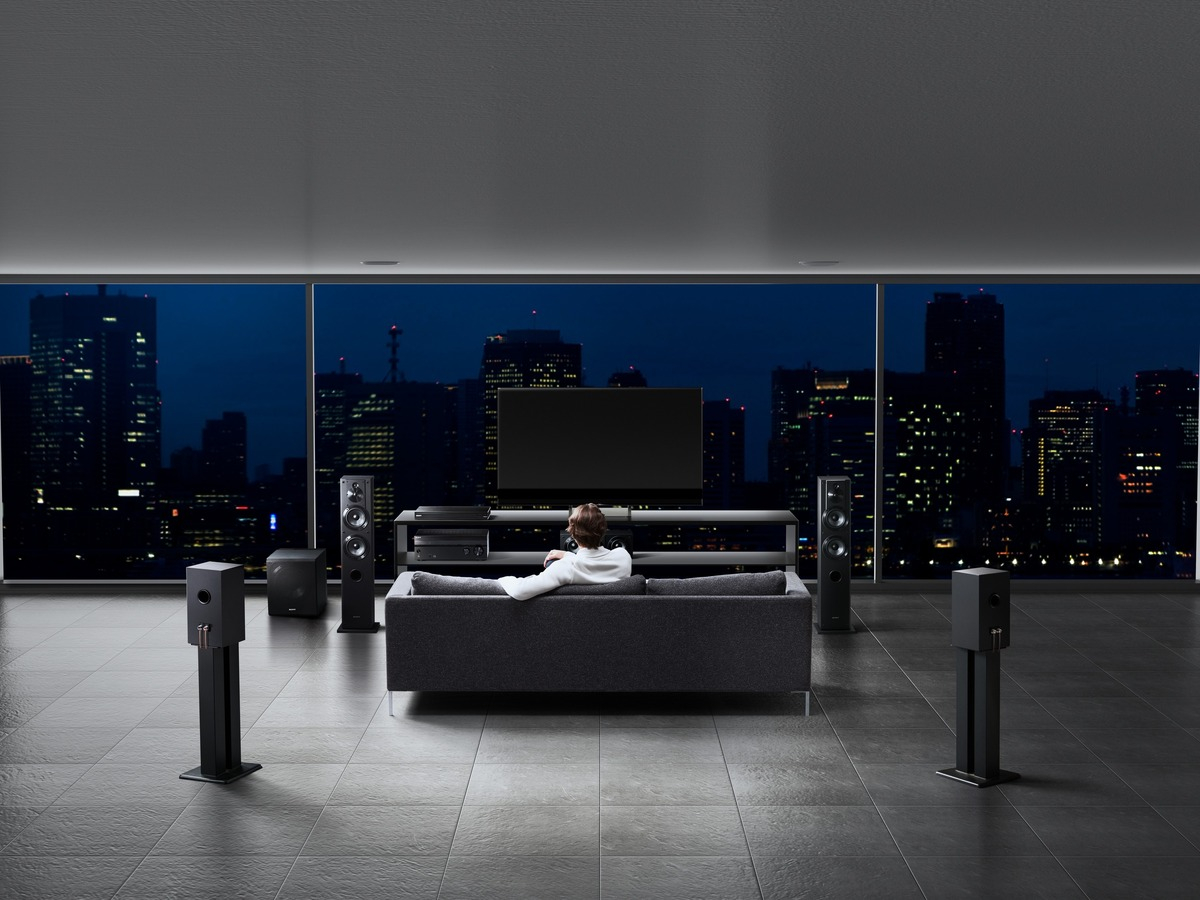 Wohnzimmer bei Nacht. Person auf einem grauen Sofa. Fernseher, Lautsprecher und Stadtansicht im Hintergrund.