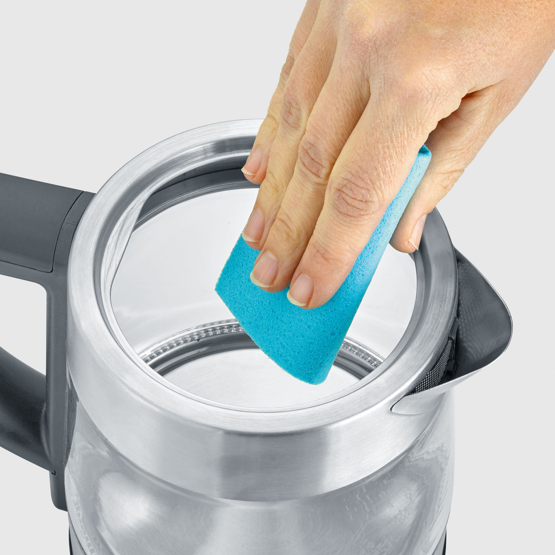 Een hand met een blauwe spons reinigt de binnenkant van een glazen en stalen waterkoker.