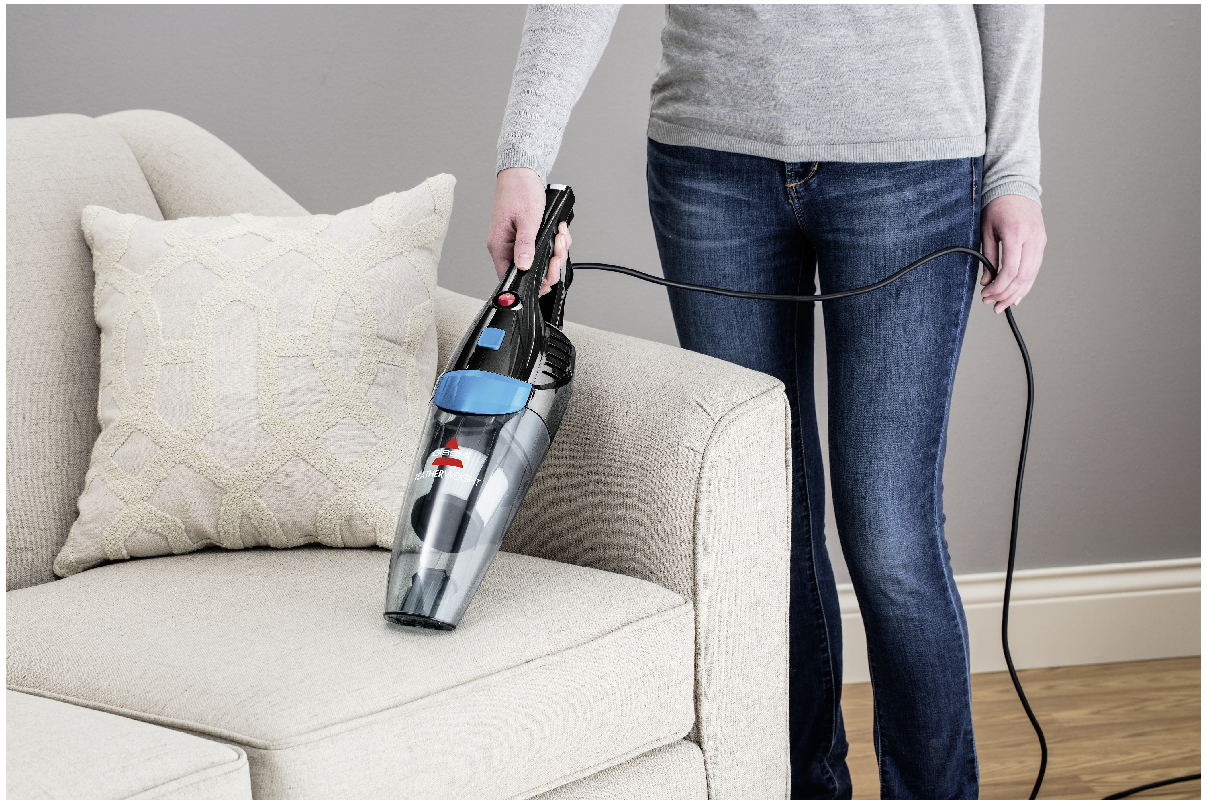 Eine Frau benutzt einen schwarz-blauen Handstaubsauger auf einem beigen Sofa.