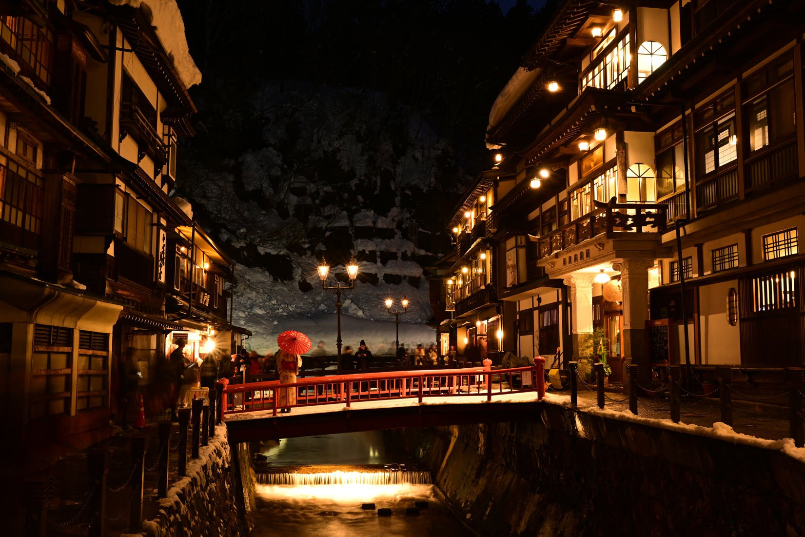 Scène nocturne d'une rue japonaise. Bâtiments éclairés, personnes, un pont rouge et un ruisseau.