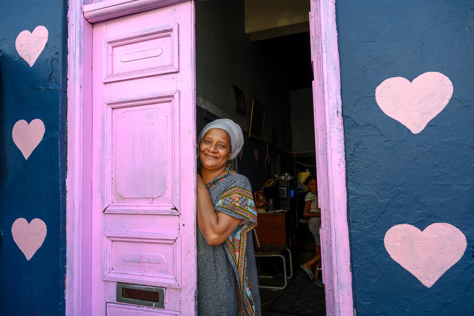 Femme dans l'embrasure, porte rose, mur bleu avec coeurs. À l'intérieur, une autre personne est visible.