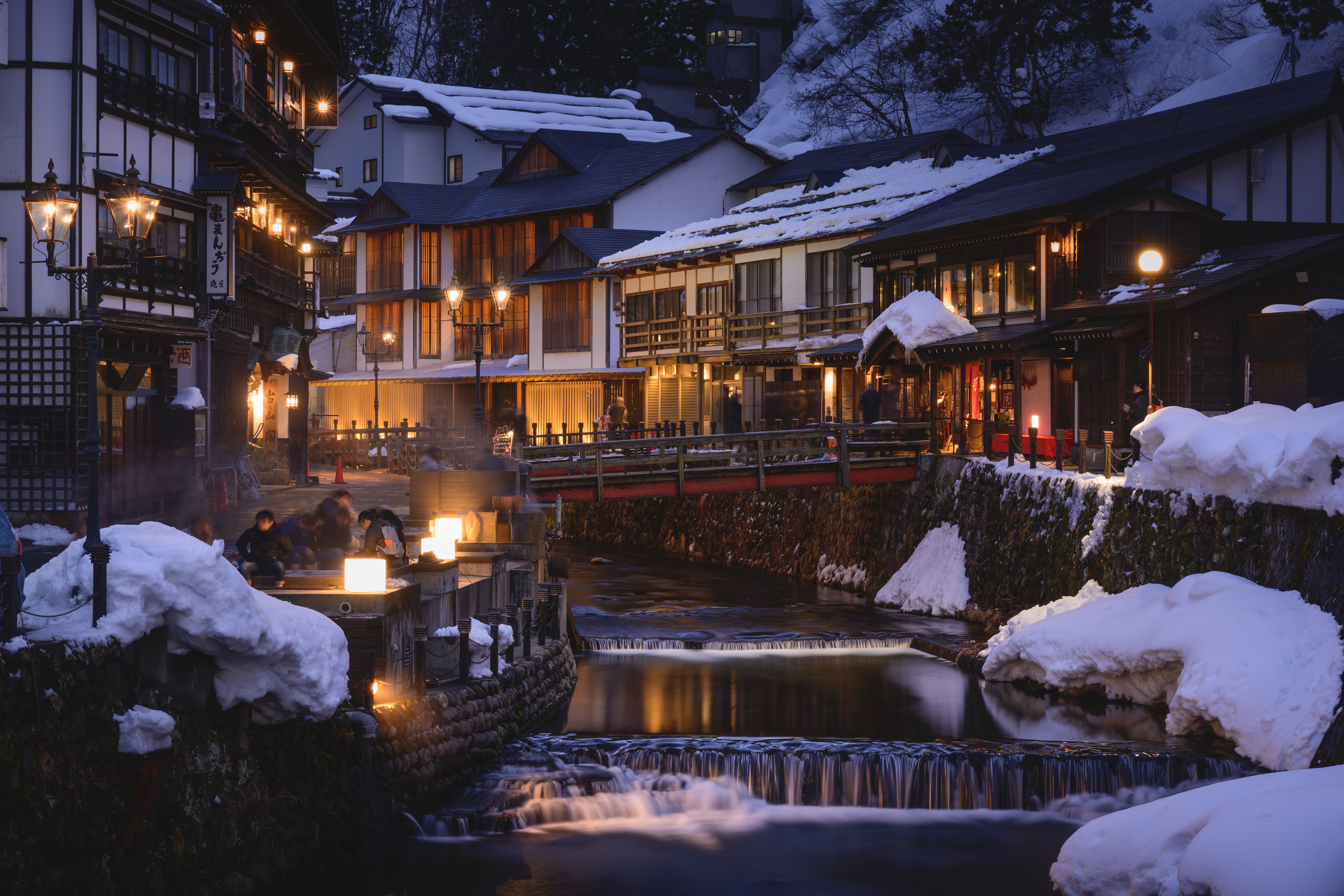 Scène de nuit d'un village japonais avec neige, lumières et une rivière.