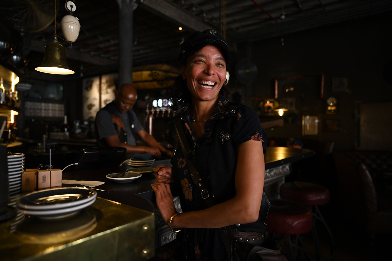 Femme souriante dans un bar. Une personne se trouve derrière le bar. Intérieur sombre, avec lumières et décor.