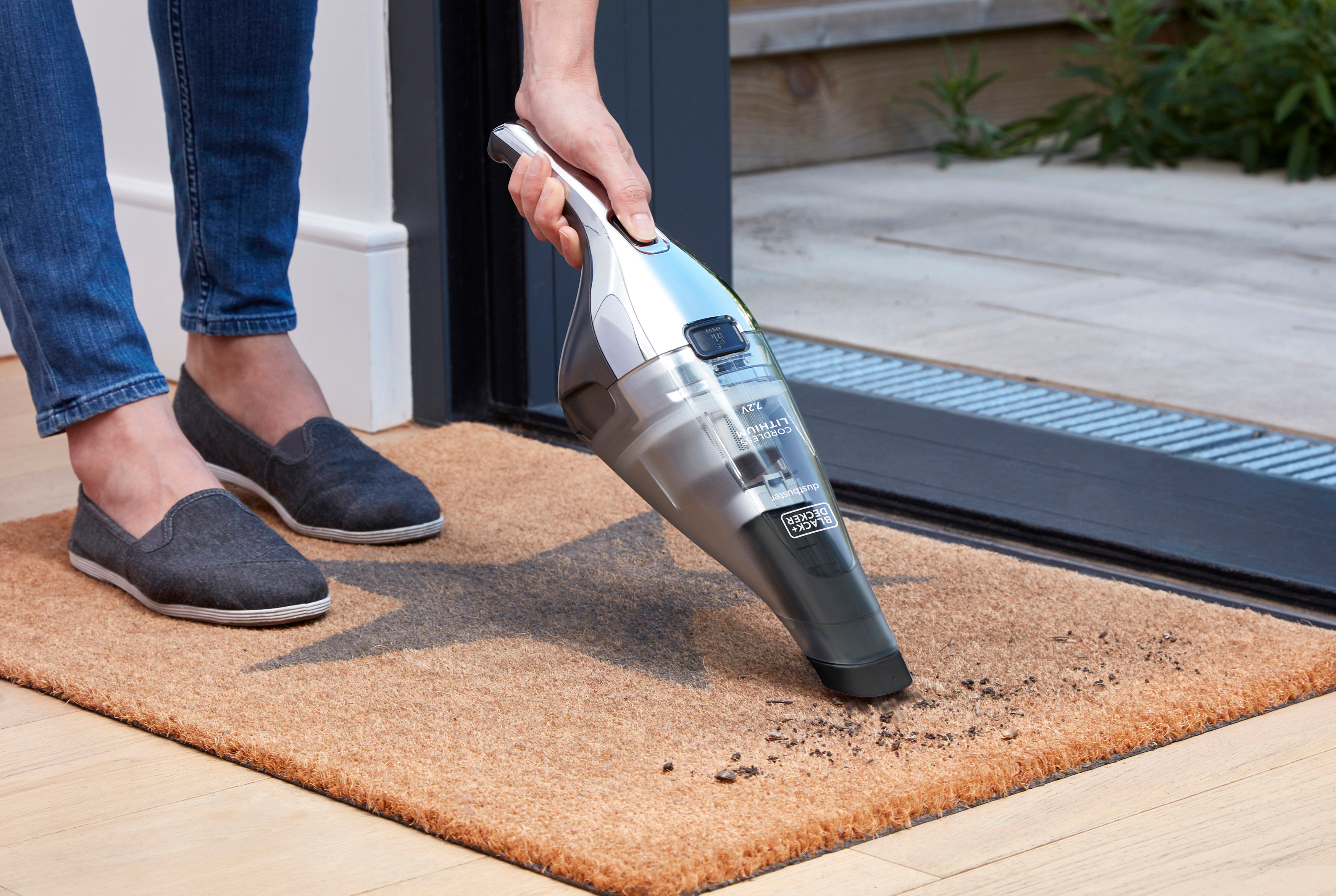 Person saugt eine Fußmatte mit einem kabellosen Staubsauger; Person trägt Jeans und graue Schuhe.