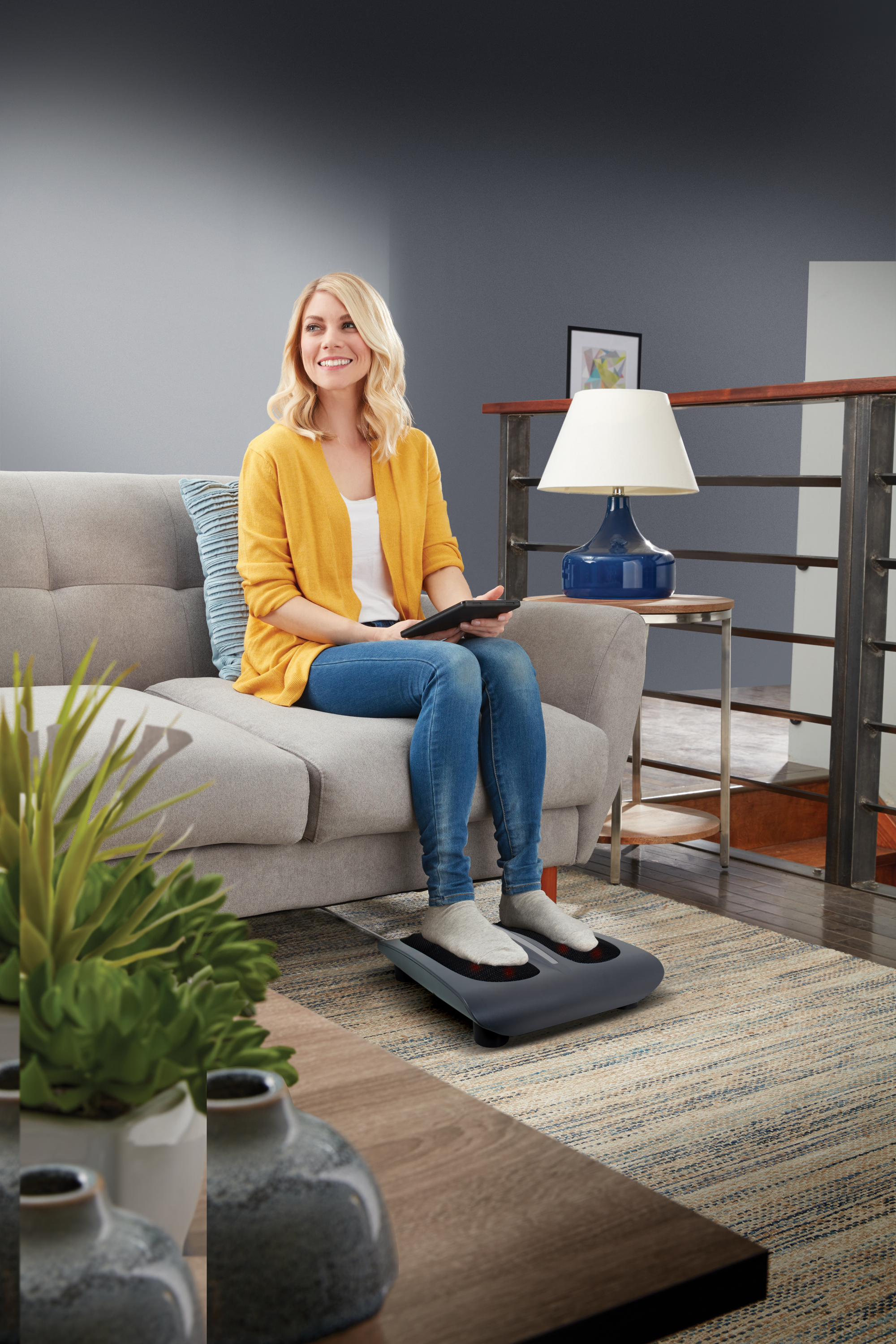 Femme assise sur un canapé gris utilisant un masseur de pieds, avec une table d'appoint et une lampe. Fond neutre.