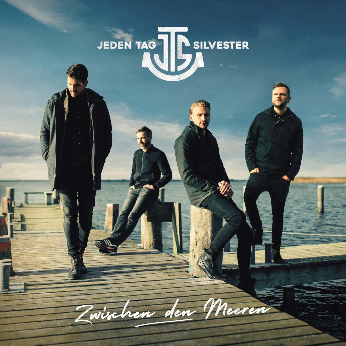 Band von vier Männern auf einem Steg, mit See und Himmel im Hintergrund.