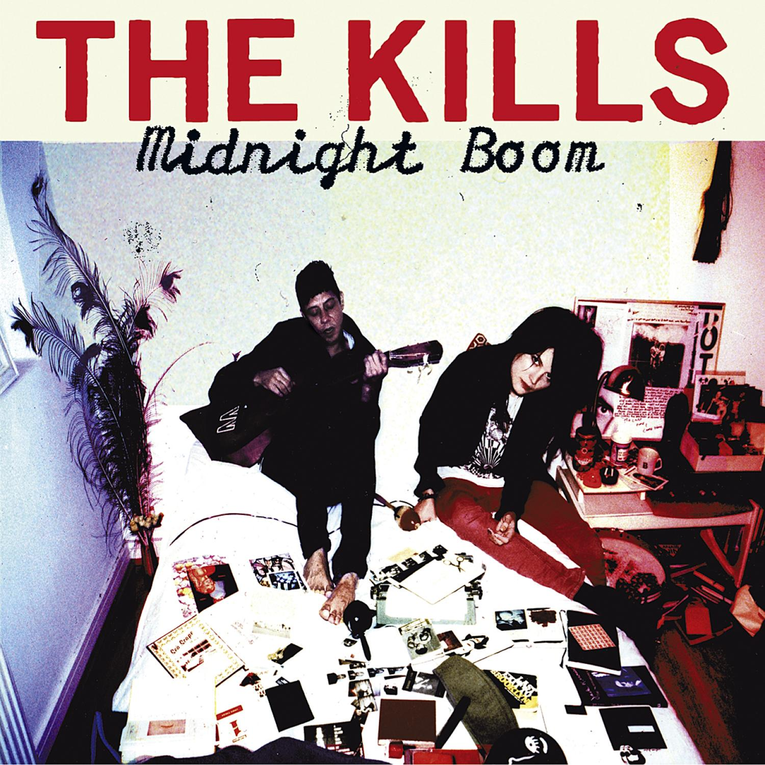Albumhoes voor The Kills' Midnight Boom. Twee mensen zitten op een bed omringd door items, met een gitaar.