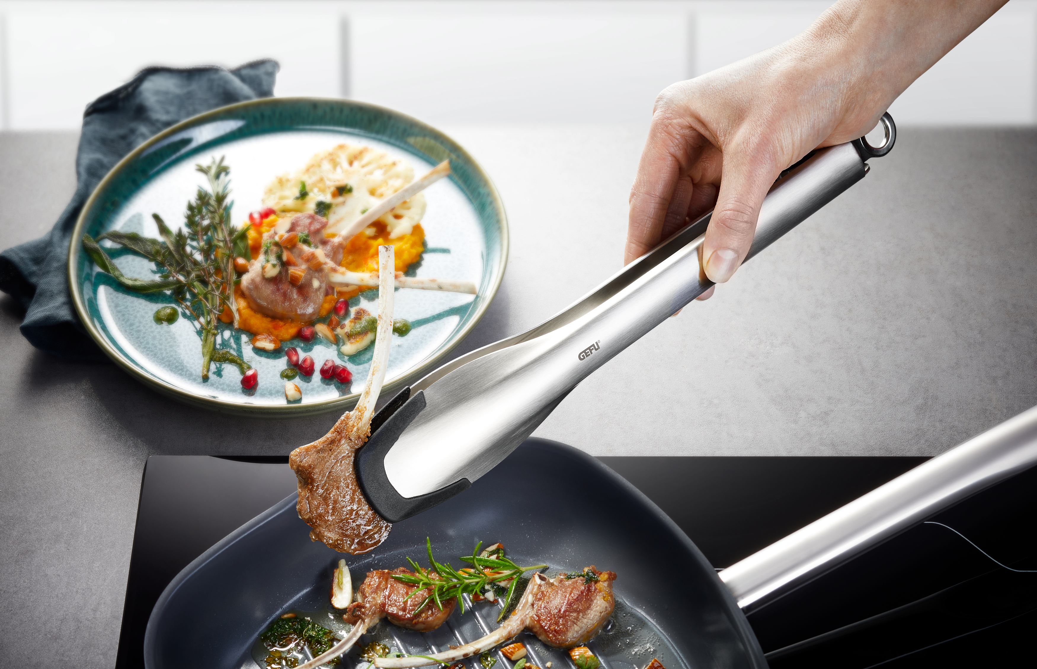 Eine Hand hebt mit einer Zange ein Lammkotelett aus einer Pfanne. Im Hintergrund ein Teller mit Lammfleisch und Beilagen.