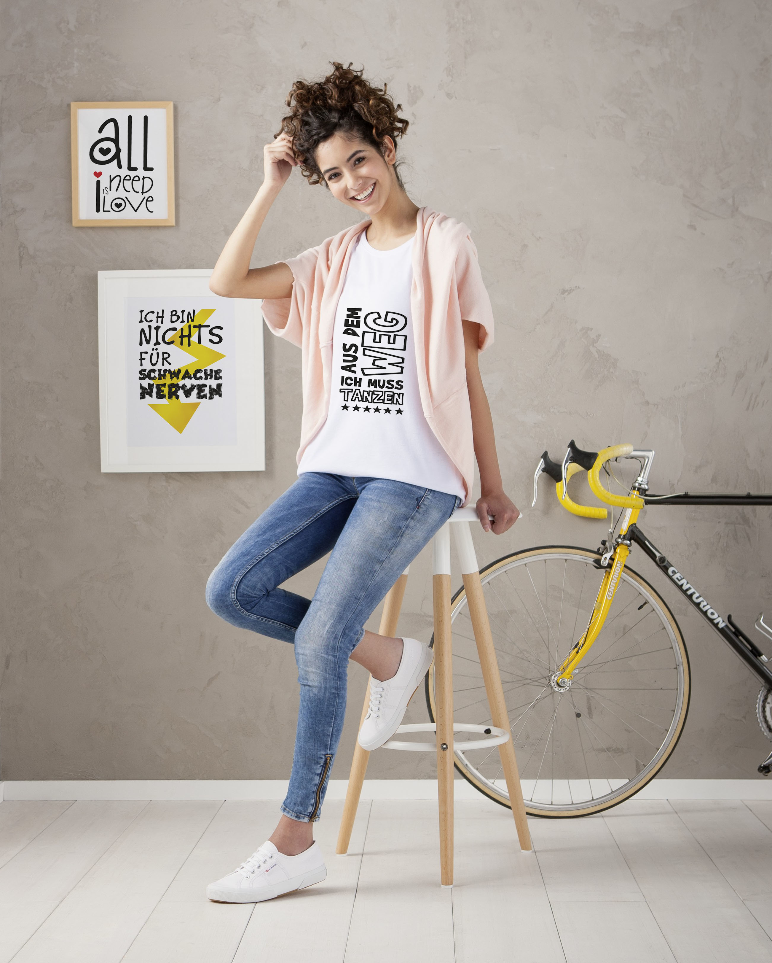 Frau sitzt auf einem Hocker, trägt ein weißes T-Shirt und Jeans. Ein Fahrrad und Kunst dahinter.