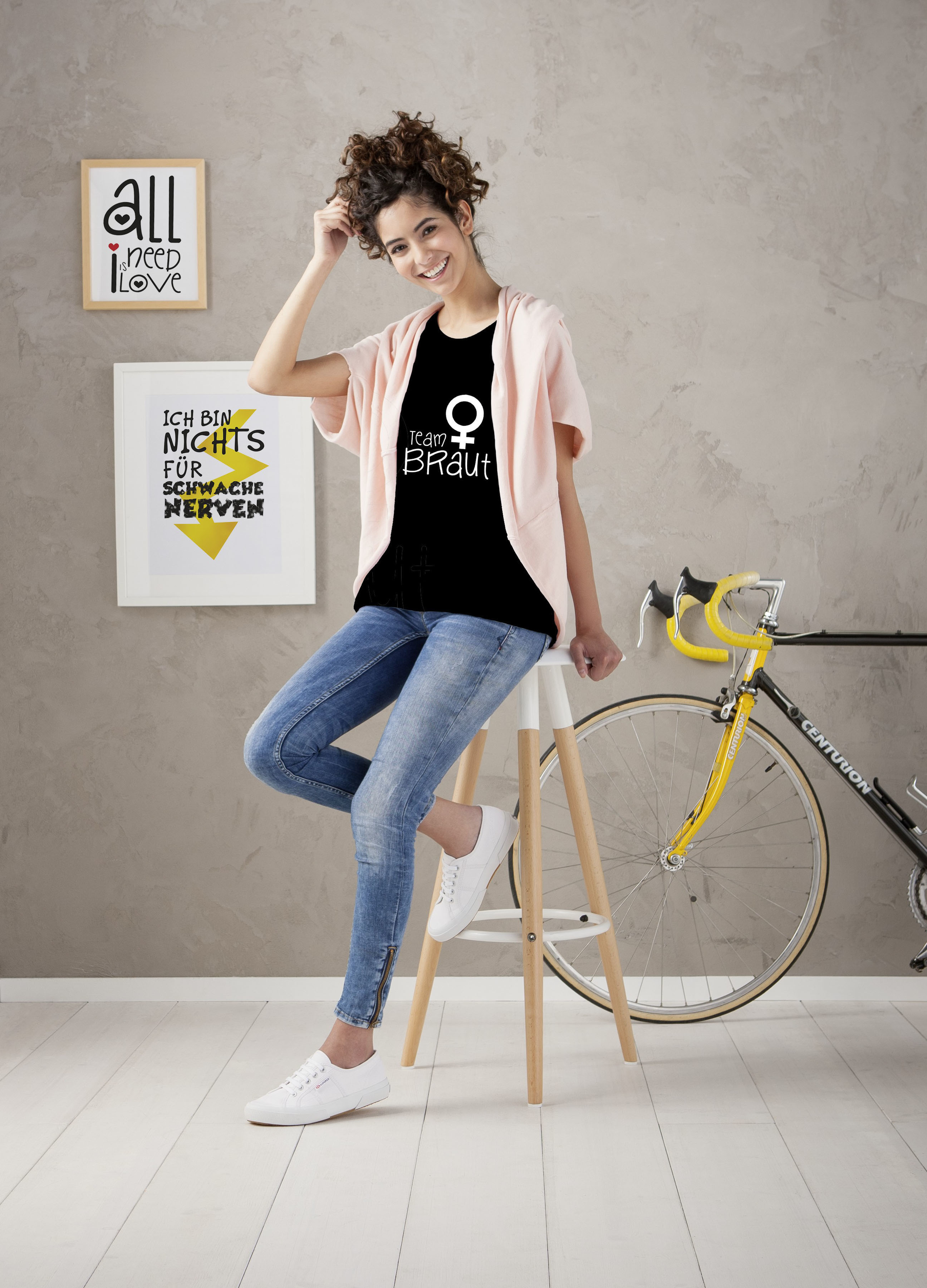 Eine Frau in Jeans und einem Shirt mit der Aufschrift "Team Braut" sitzt auf einem Hocker.