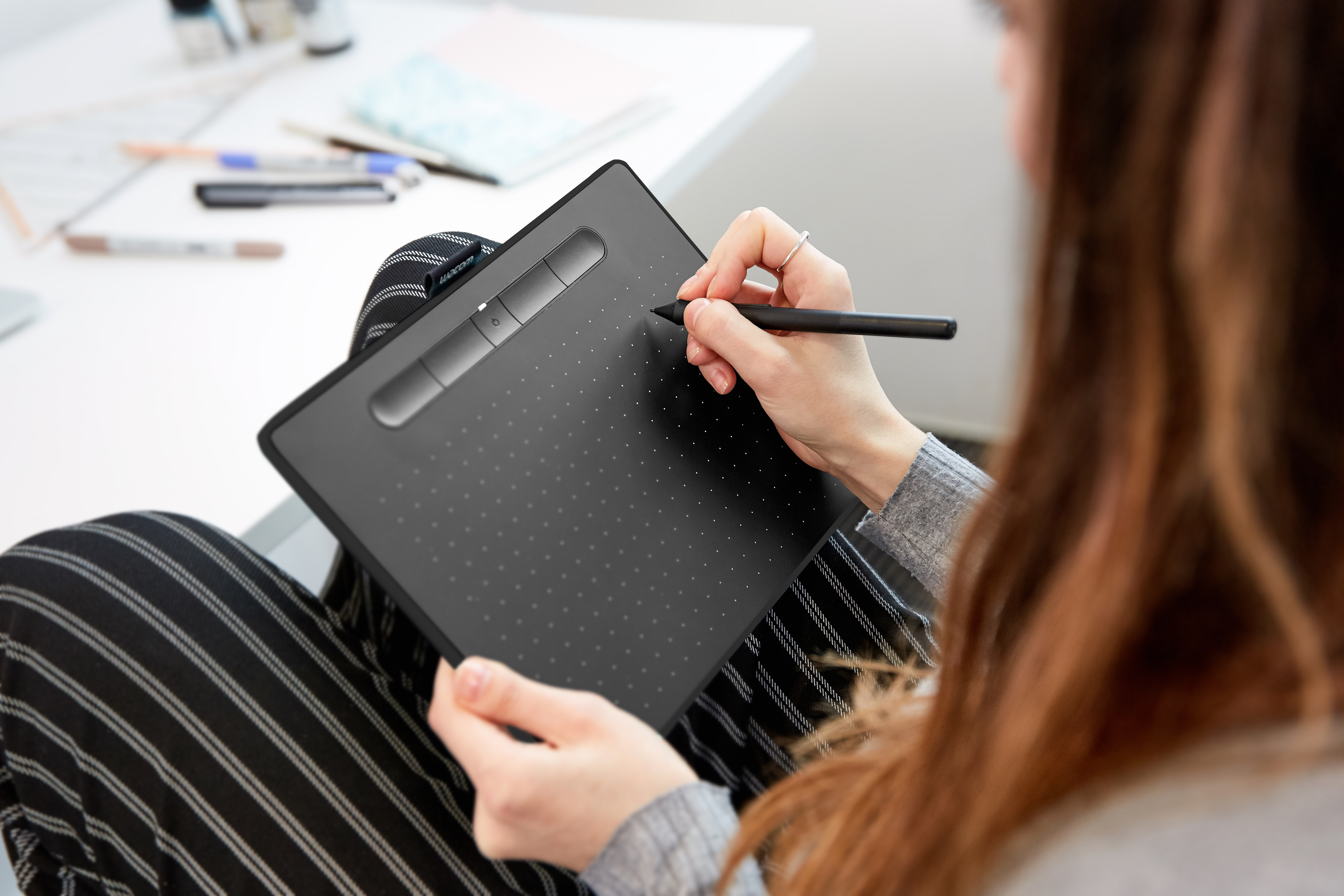 Eine Frau benutzt ein schwarzes Grafiktablett mit Stift. Sie sitzt. Der Hintergrund ist unscharf.