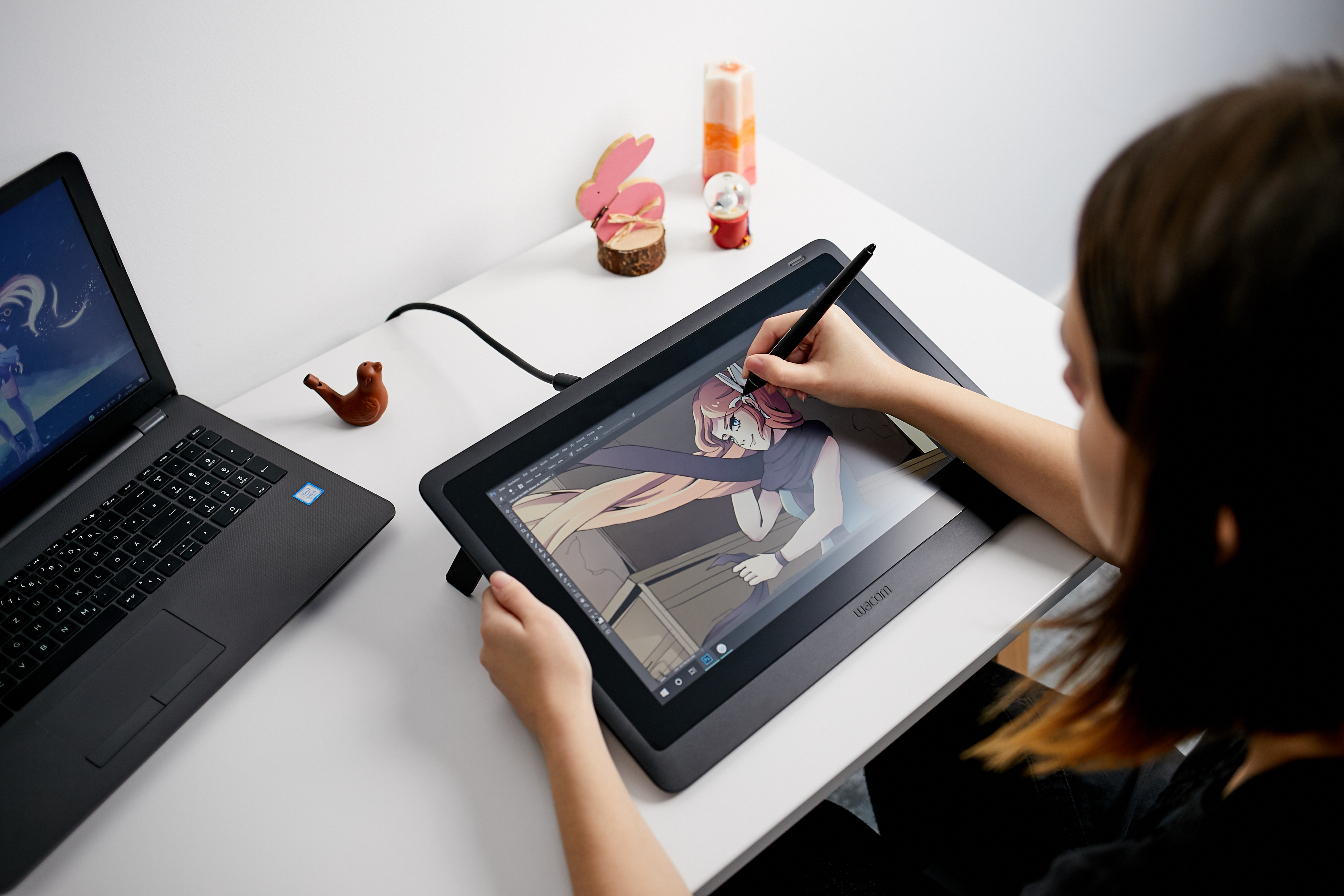 Eine Person zeichnet mit einem Stift auf einem Wacom-Tablet. Ein Laptop und Dekorationen stehen auf dem weißen Schreibtisch.