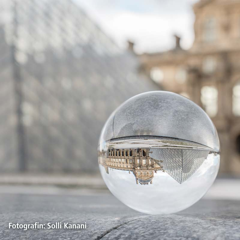Egy kristálygömb a Louvre Múzeumot és egy üvegpiramist tükröz. A háttérben elmosódott épületek láthatók.