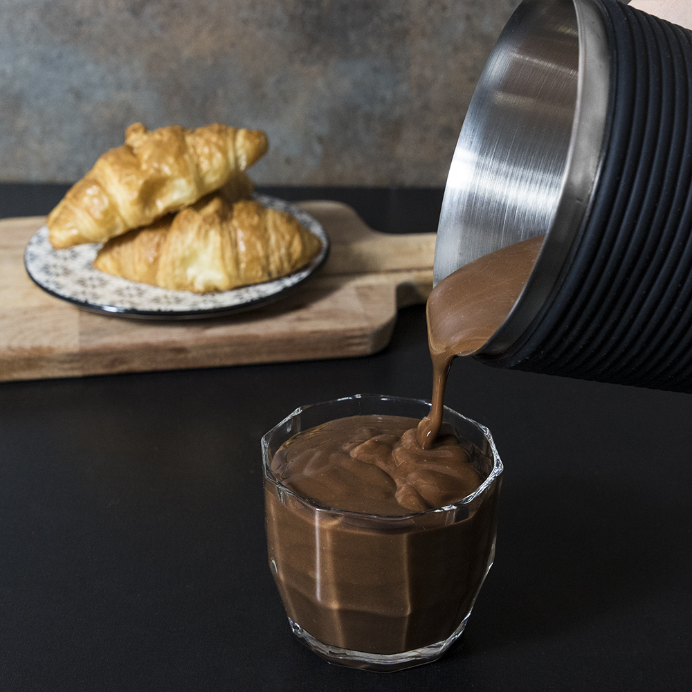 Chocolate vertiéndose en un vaso con cruasanes en una tabla de madera en el fondo.