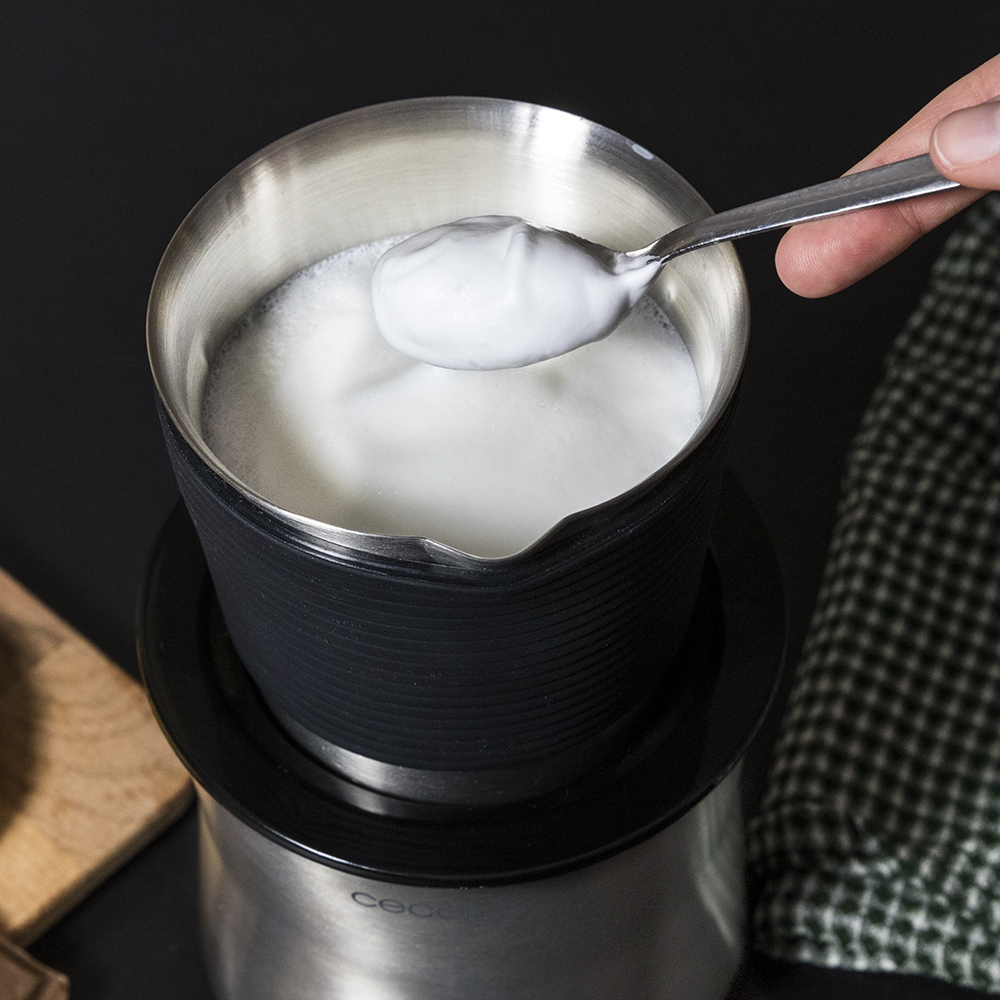 Una cuchara saca espuma de una jarra espumadora de acero inoxidable sobre una base negra, con un paño verde cerca.