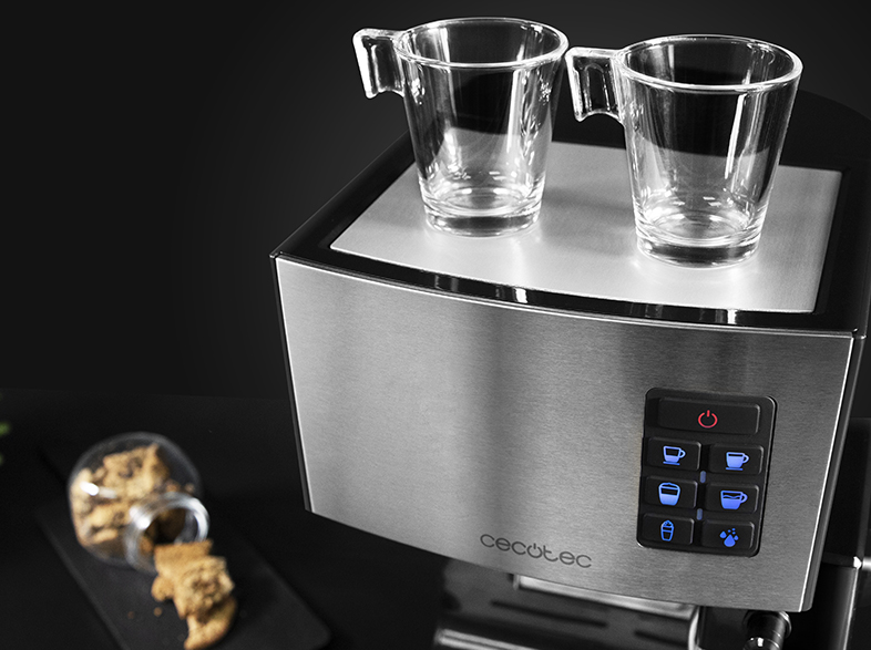 Una cafetera de acero inoxidable con dos vasos de vidrio encima. Galletas en una superficie negra en primer plano.
