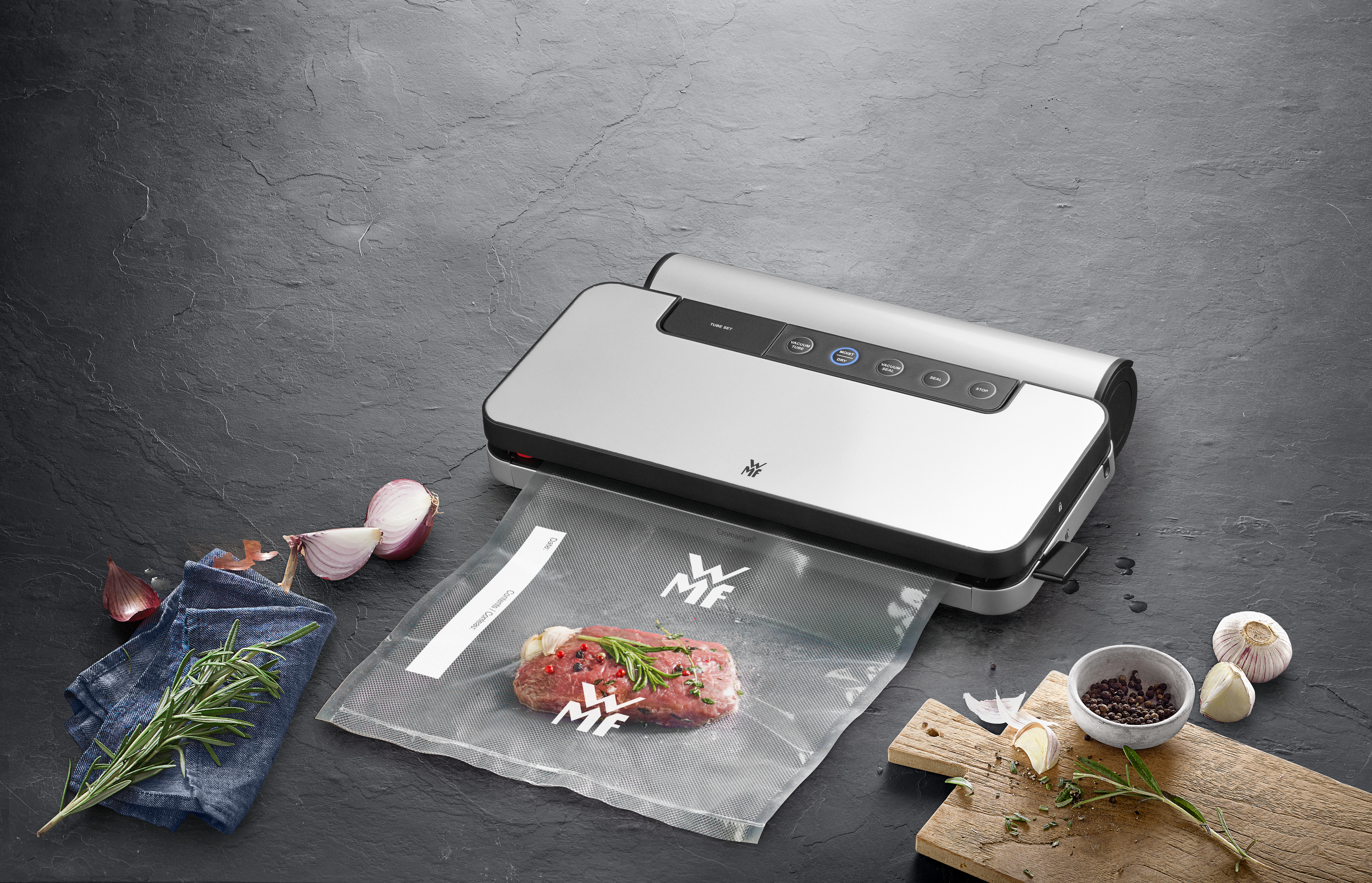 Eine Vakuumversiegelungsmaschine mit Fleisch in einem Beutel und Zutaten auf dunkler Oberfläche.