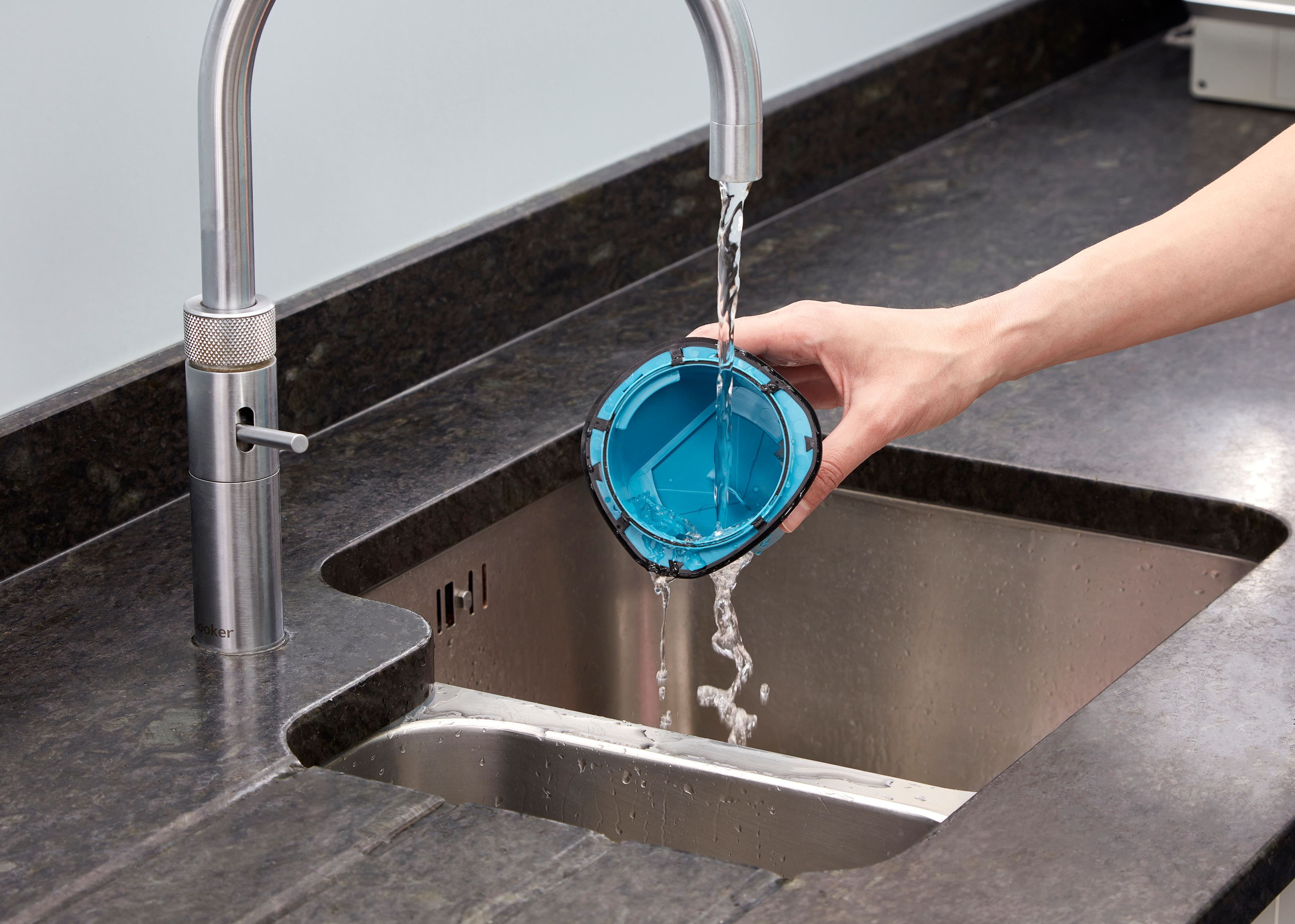 Une main rince une tasse filtrante bleue sous un robinet chromé dans un évier en acier inoxydable.