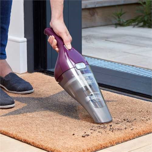A person vacuuming a brown mat with a purple and gray handheld vacuum cleaner.