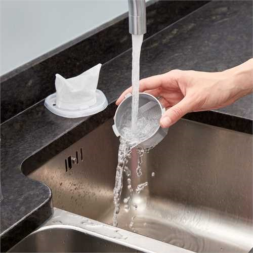 Person rinsing a filter under a faucet in a stainless steel kitchen sink.