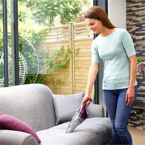 Woman cleaning a gray couch with a handheld vacuum. She wears a light blue shirt and jeans.