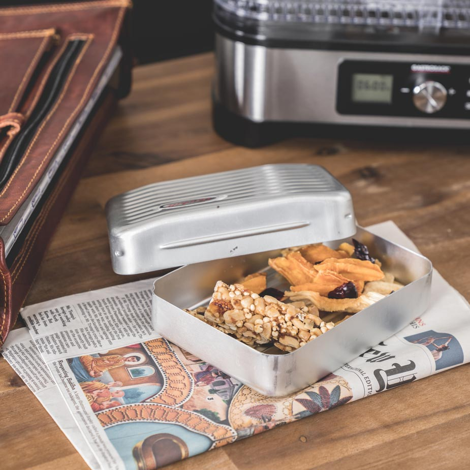 Eine silberne Brotdose mit Snacks und ein Trockner auf einem Holztisch mit Zeitung.