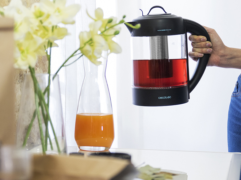Una mano sostiene una tetera. Un jarrón con flores y un vaso de jugo junto a ella.